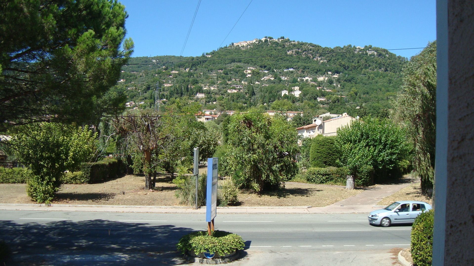 Une route avec un panneau indiquant la présence d’un hôtel. On aperçoit des arbres et, au loin, une colline avec des bâtiments.