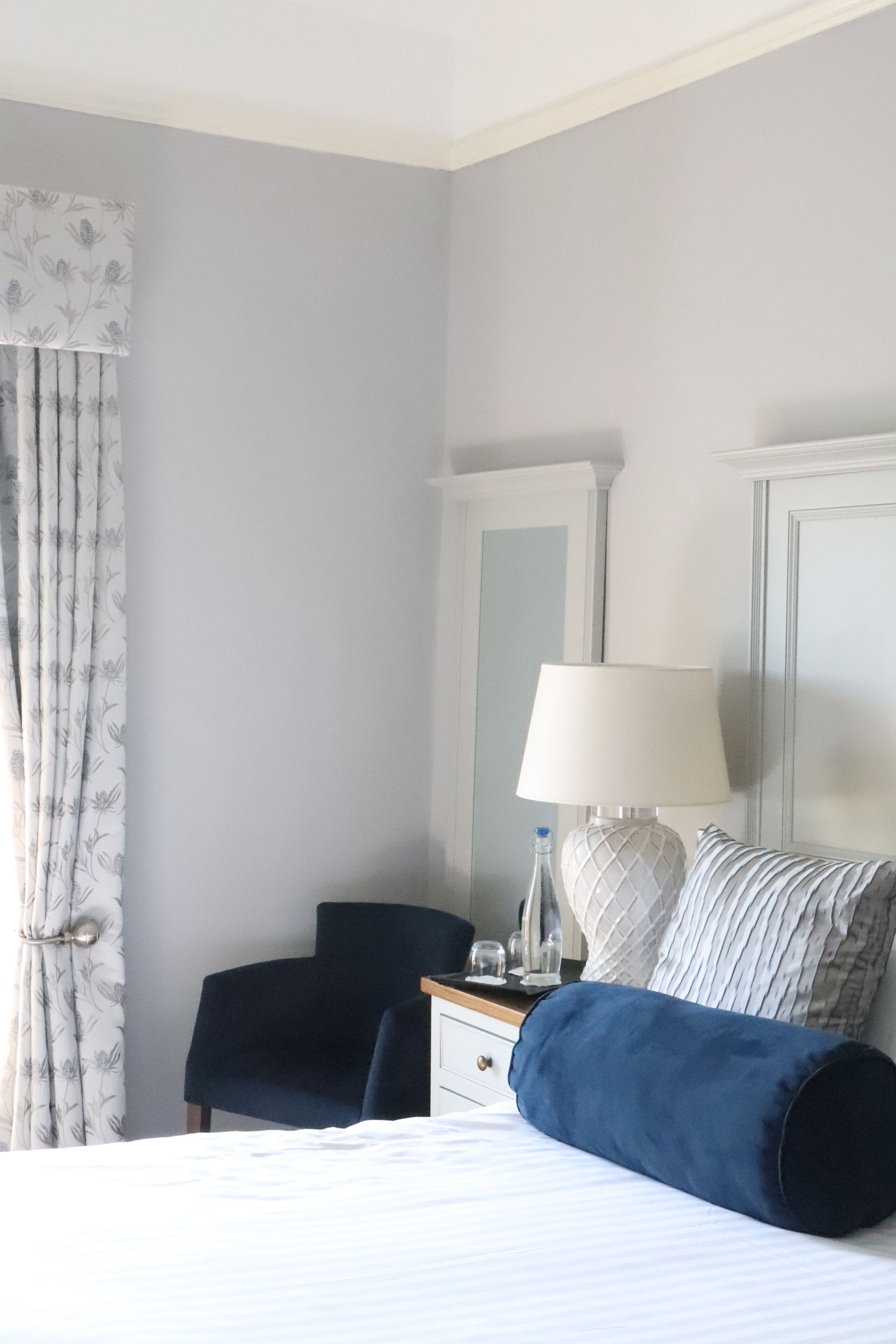 A hotel room with a bed, a desk, a chair, a lamp, and a window with curtains.