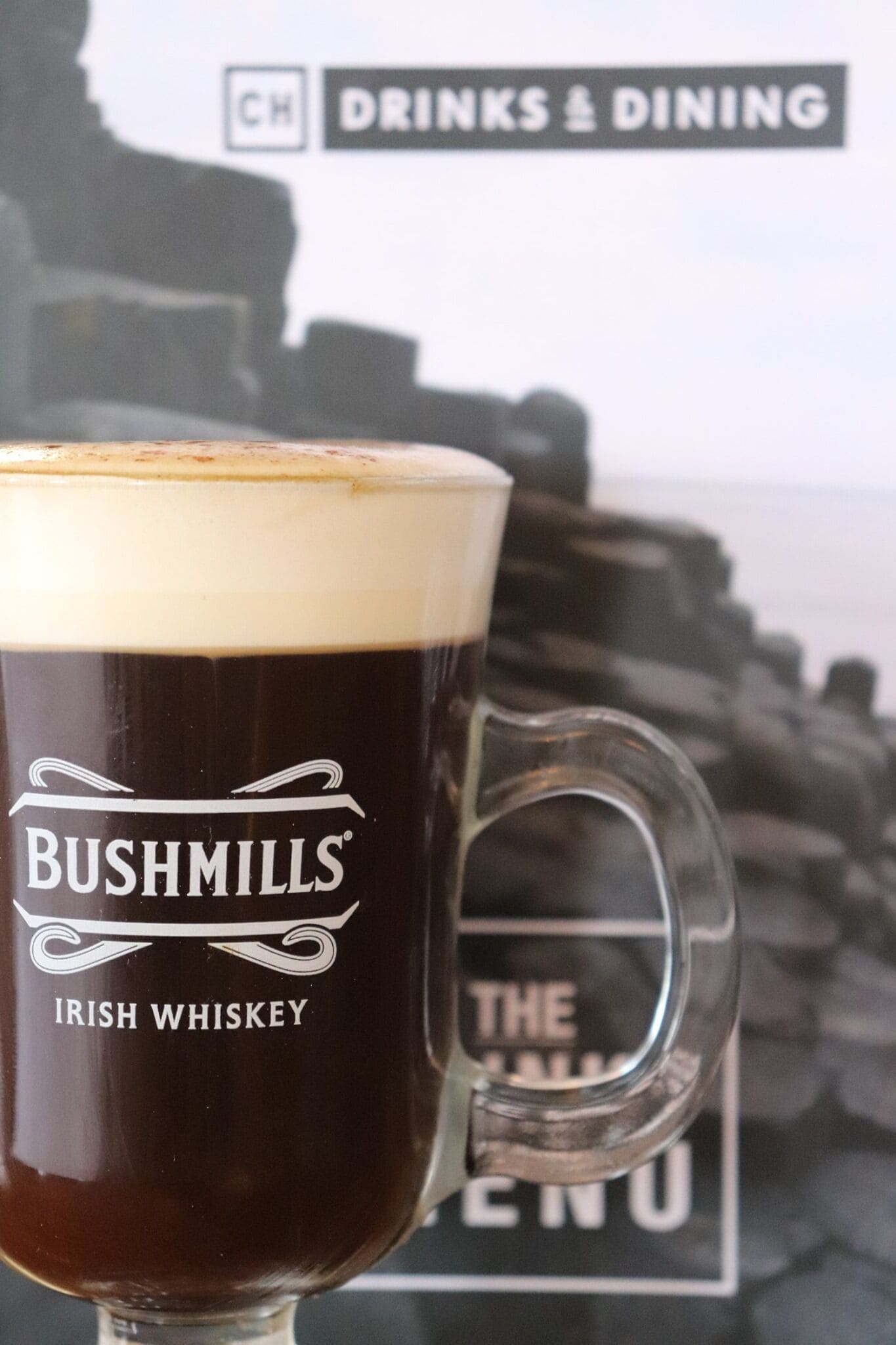 A glass of Irish whiskey with a frothy head, labeled "BUSHMILLS IRISH WHISKEY," in front of a menu board with "DRINKS & DINING" visible.