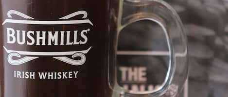 A glass of Irish whiskey with a frothy head, labeled "BUSHMILLS IRISH WHISKEY," in front of a menu board with "DRINKS & DINING" visible.