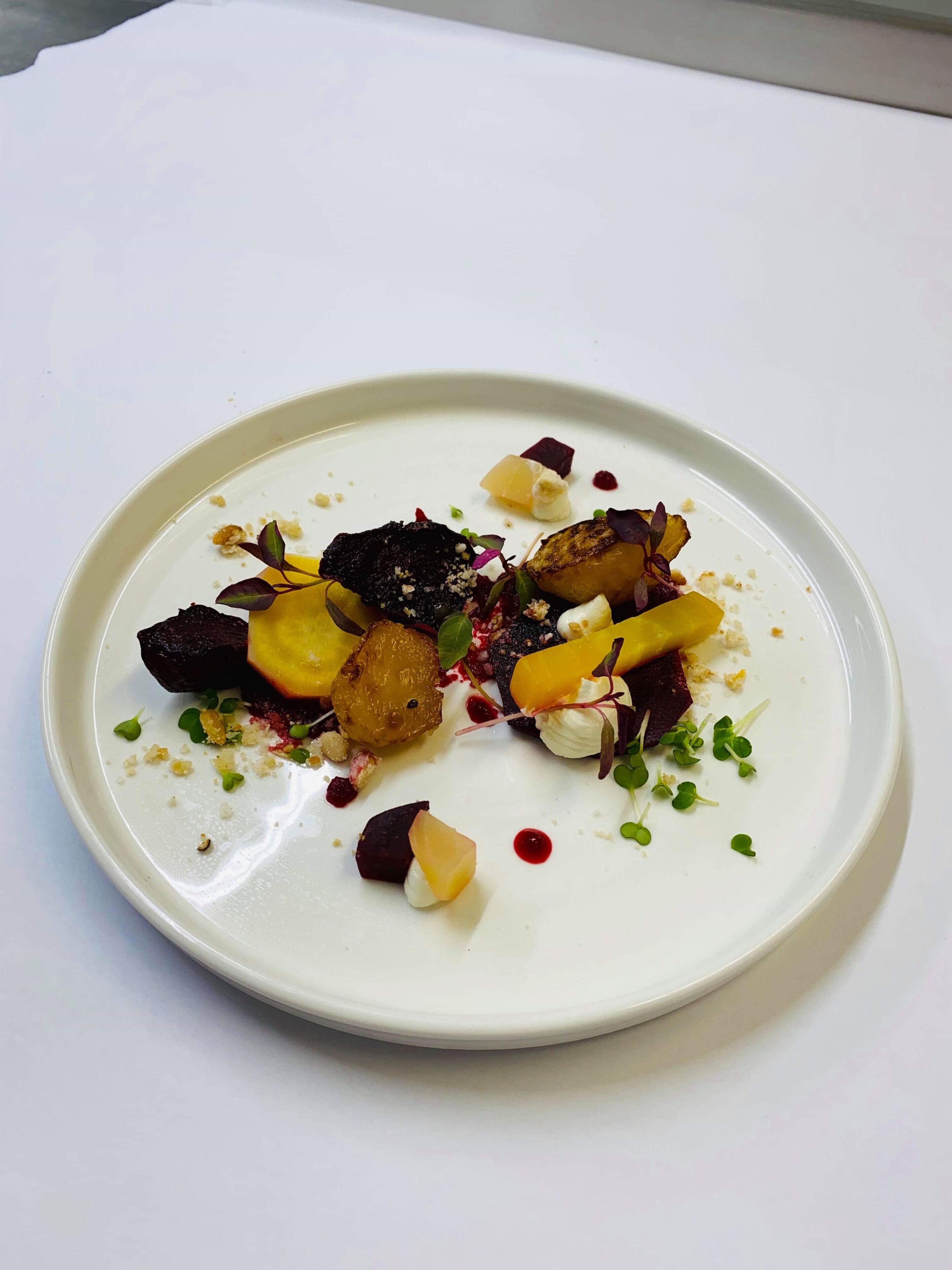 A white plate with a variety of food items including what appears to be roasted potatoes, beetroot, and possibly some greens, arranged on a white tablecloth.