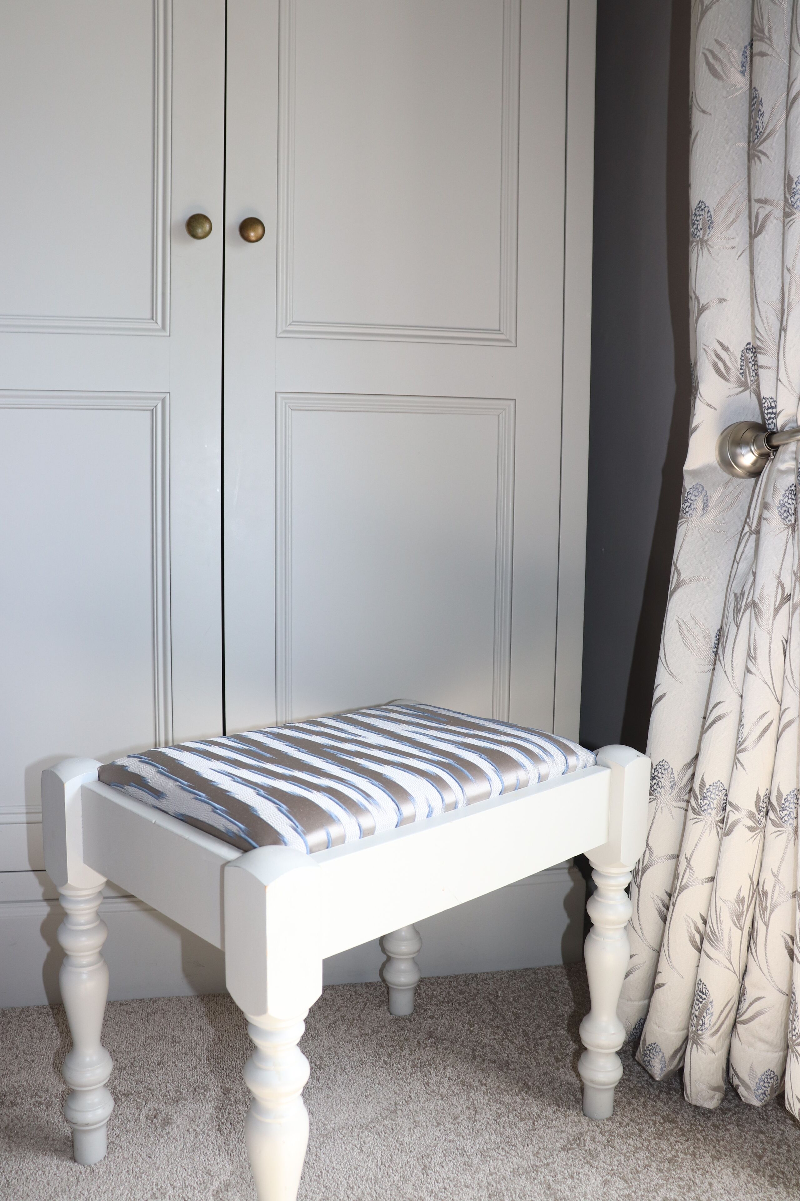 A white bench with a striped cushion in front of a white door with two brass handles.