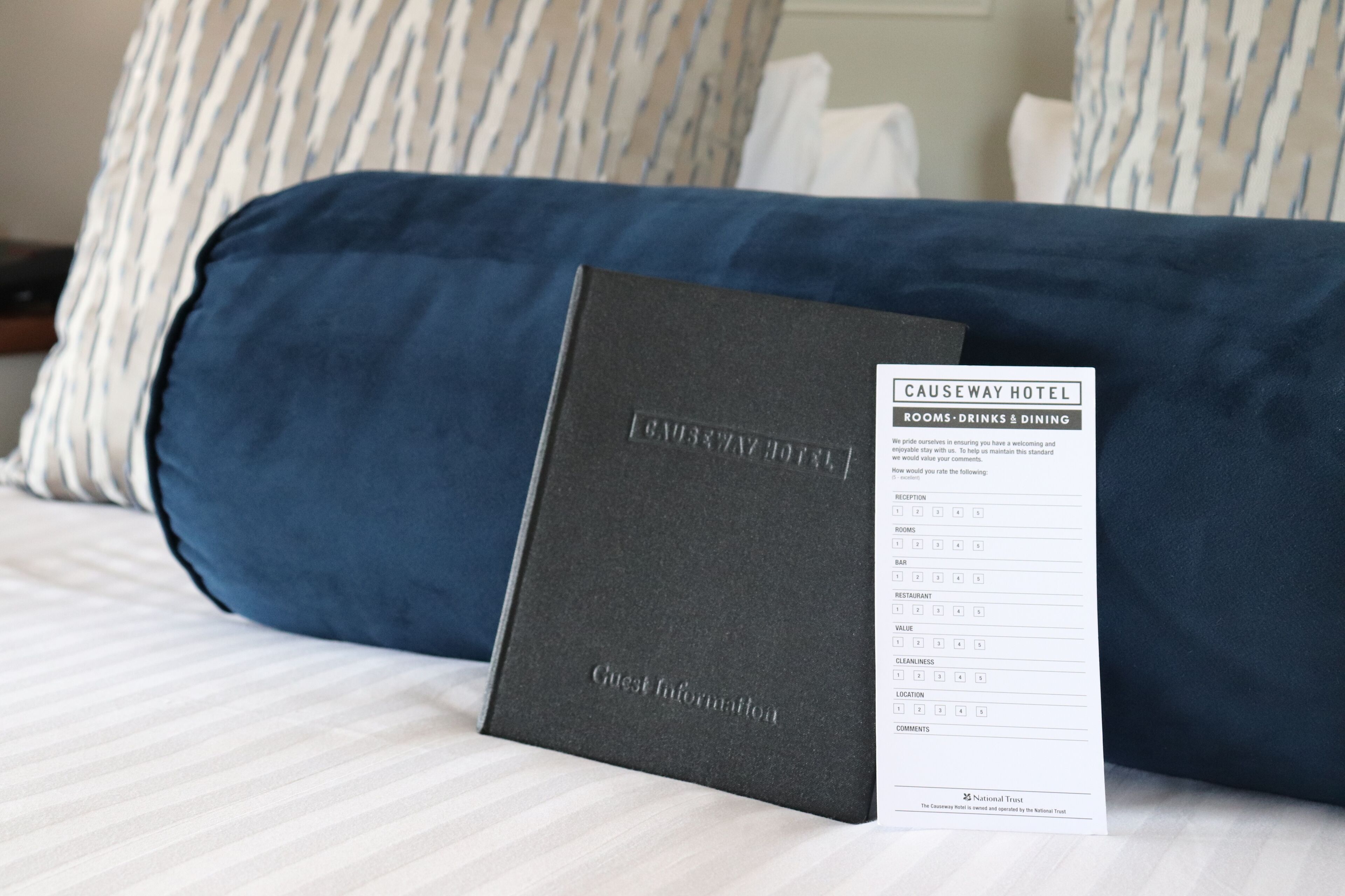 Une chambre d’hôtel avec un lit agrémenté d’oreillers à rayures, une couvre-lit bleu foncé et un petit livret noir intitulé « Informations pour les clients » posé sur le lit.