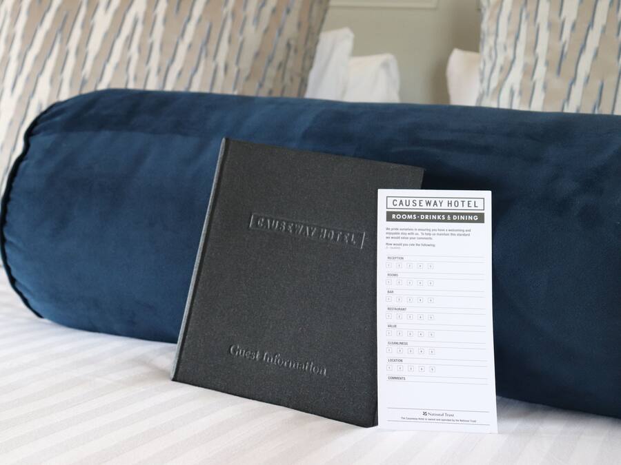 Une chambre d’hôtel avec un lit agrémenté d’oreillers à rayures, une couvre-lit bleu foncé et un petit livret noir intitulé « Informations pour les clients » posé sur le lit.
