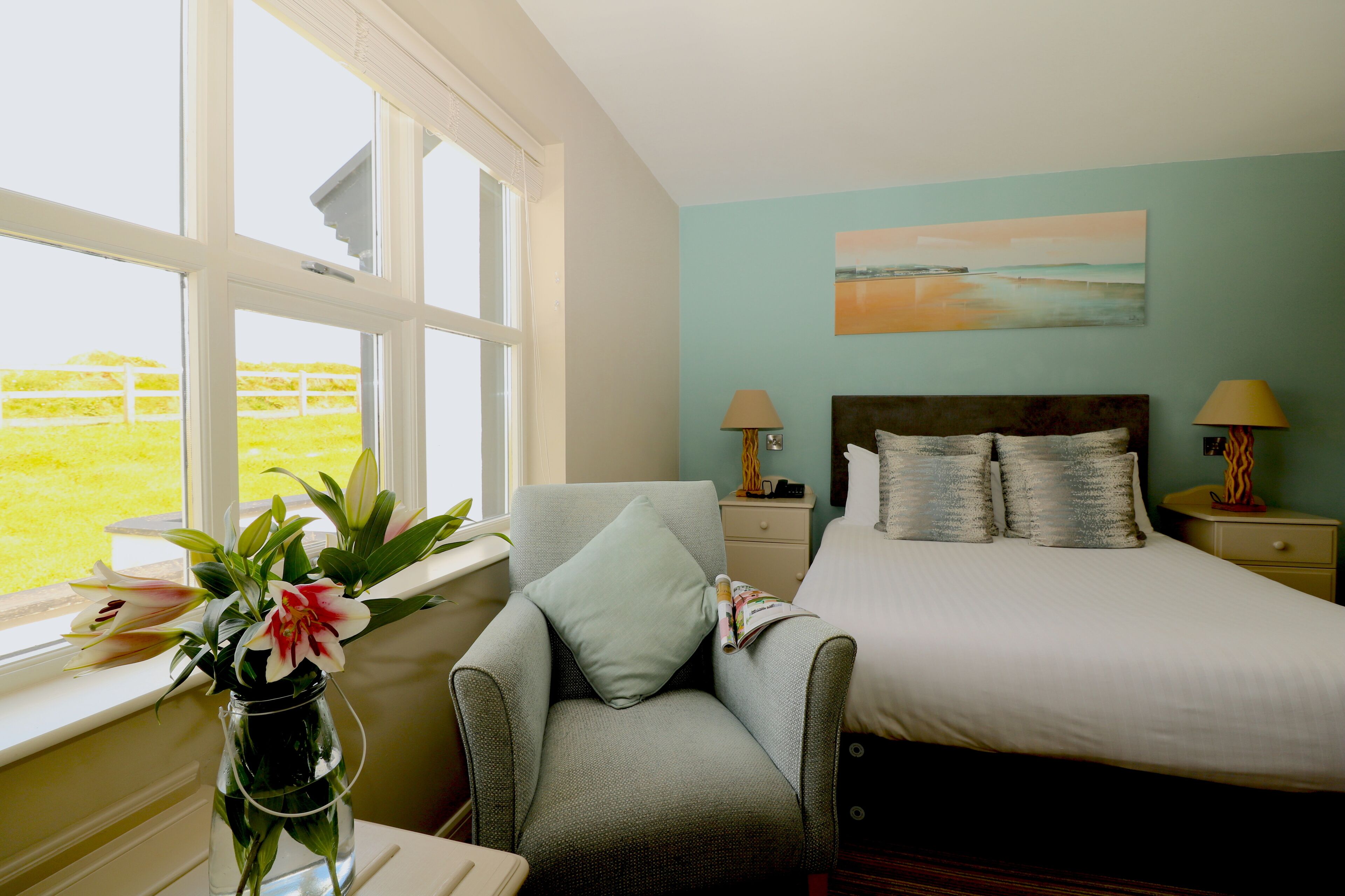 Une chambre à coucher comprenant un lit, une chaise, une table de chevet, une lampe, un vase avec des fleurs et une fenêtre donnant sur un champ verdoyant.