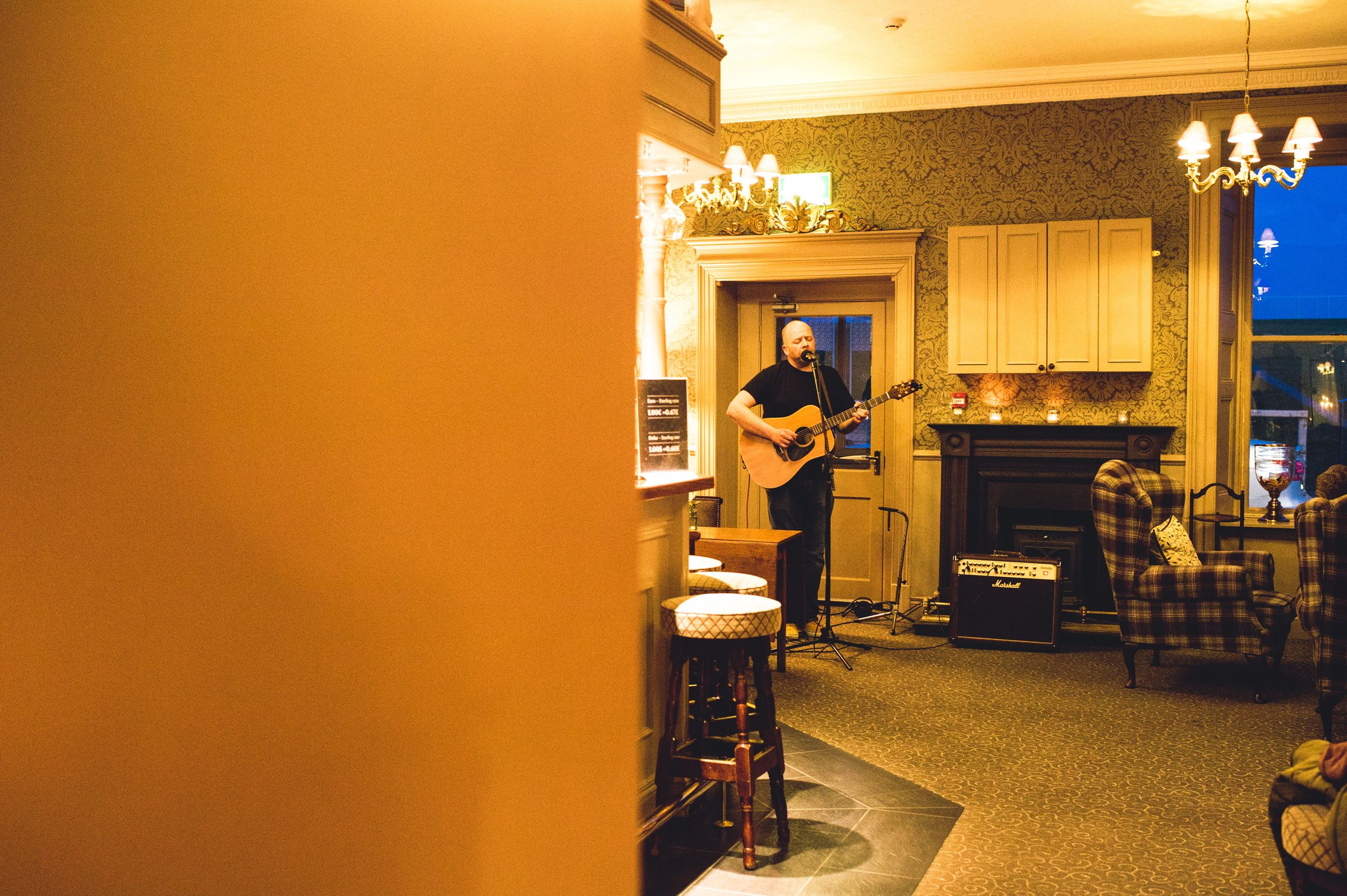 A man playing guitar in a warmly lit room with a fireplace, bar stools, and a patterned carpet.