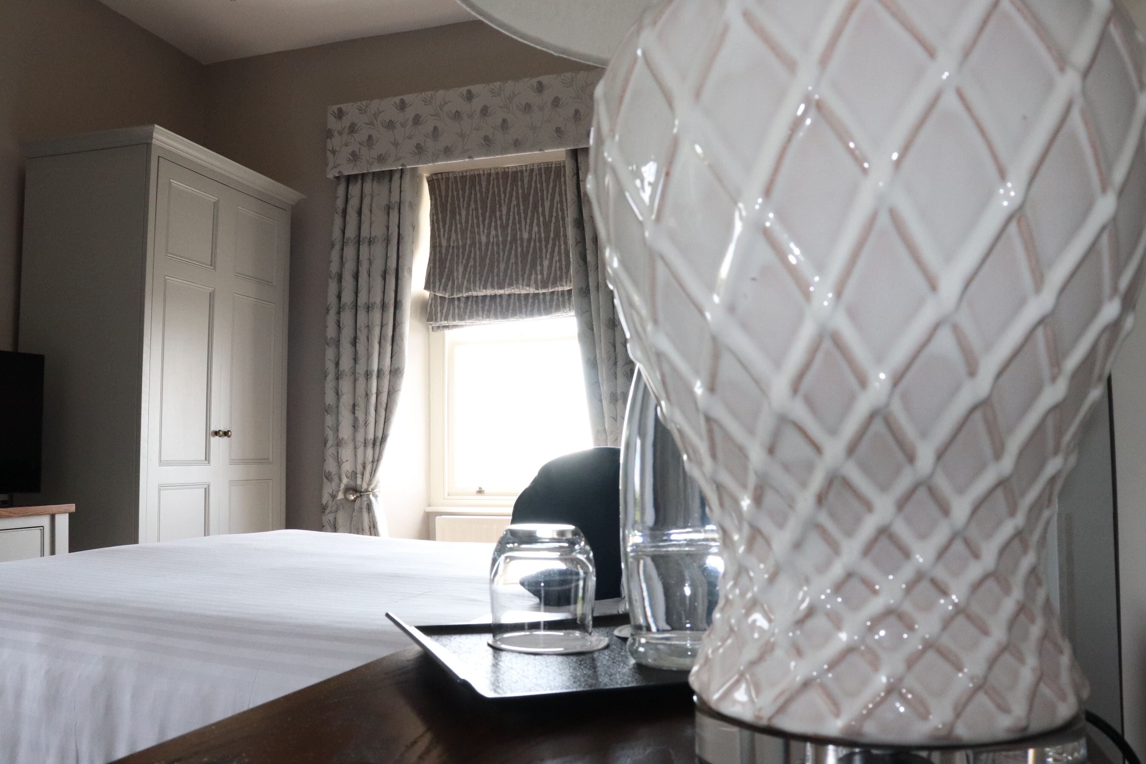 A hotel room with a bed, a bedside table with glasses and a cup, a wardrobe, and a window with curtains.