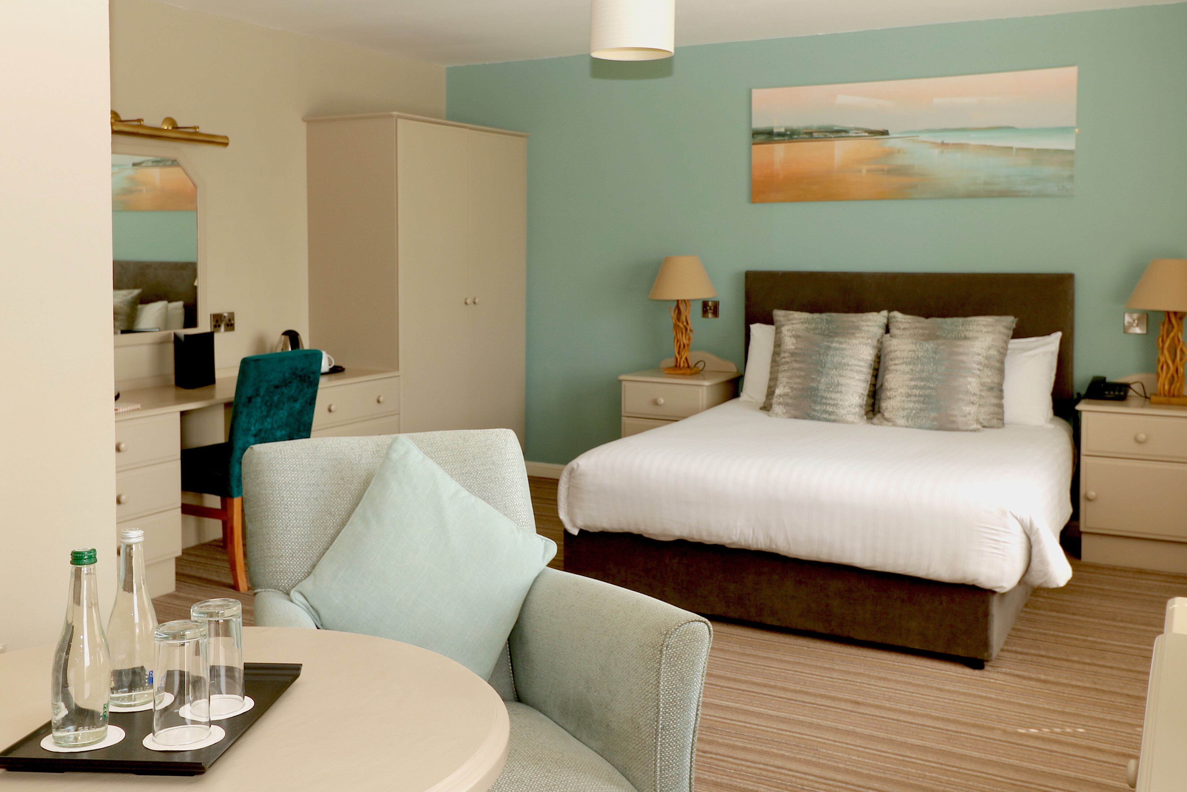A hotel room with a bed, bedside lamps, a chair, a small table with glasses, and a wardrobe.