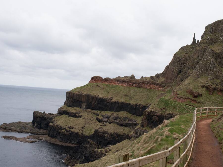 A coastal path with a wooden railing, rocky cliffs, and the sea in the background.