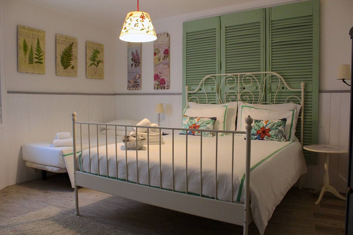 Un dormitorio con estructura de cama metálica, contraventanas verdes, una mesita de noche pequeña y una lámpara colgante.