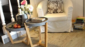Una escalera con barandillas blancas, un sillón blanco con un cojín floral, una pequeña mesa redonda con un jarrón de flores y manzanas, y una pila de libros.