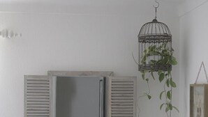 Un baño con tocador blanco, espejo con contraventanas, planta colgante y un cuadro en la pared.