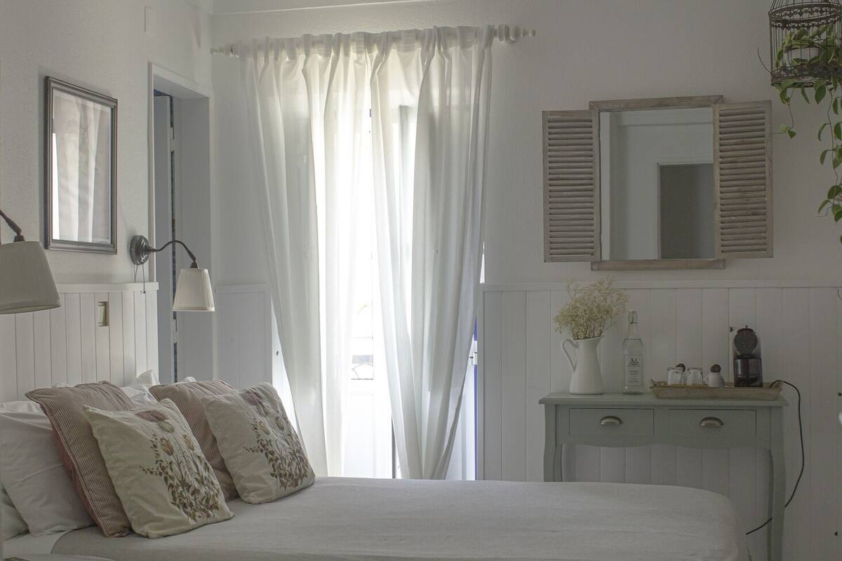 Un dormitorio con cama, mesita de noche, cómoda, espejo y ventana con cortinas.