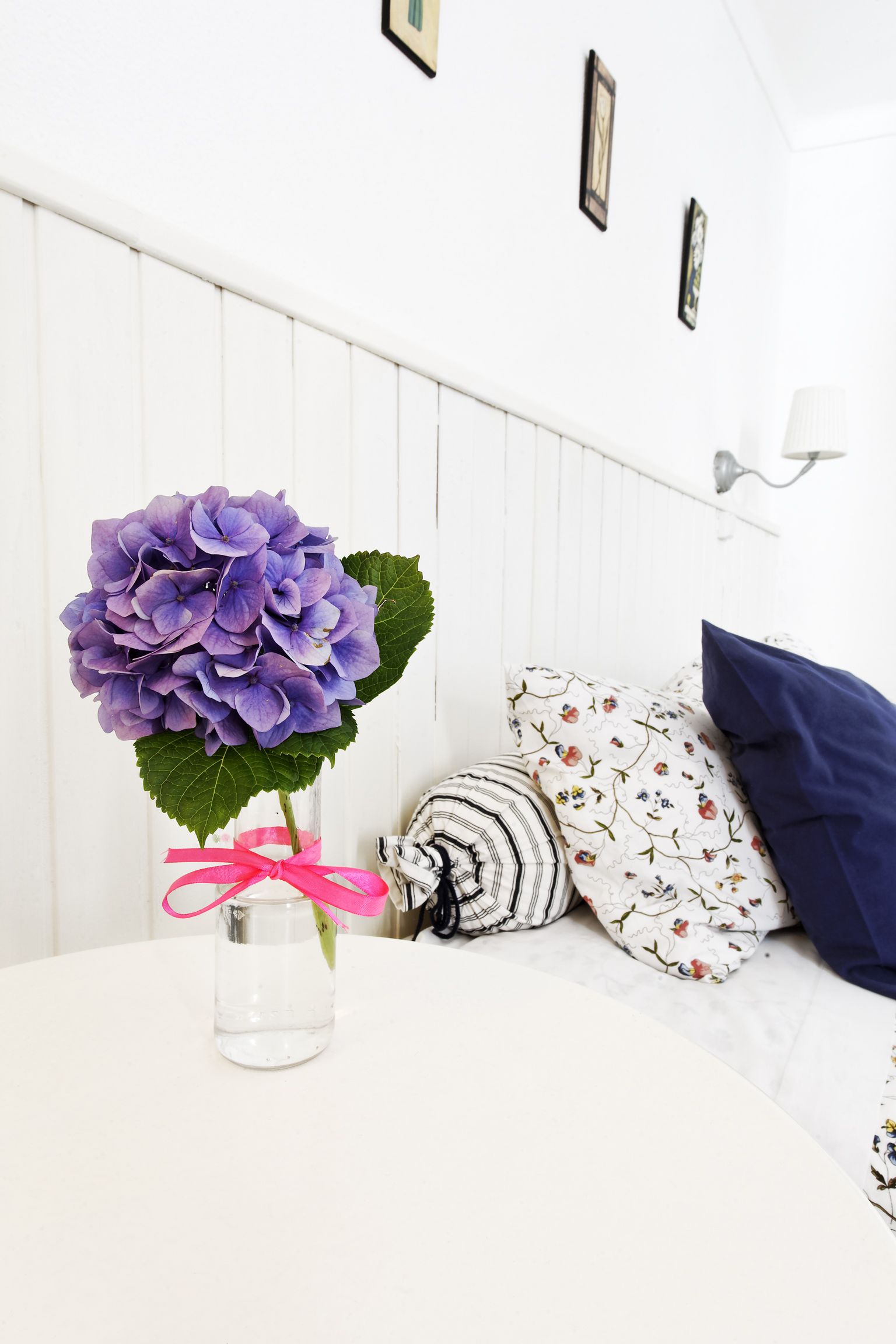 Une pièce blanche avec un lit, un vase contenant des fleurs violettes et un coussin orné d’un motif floral.