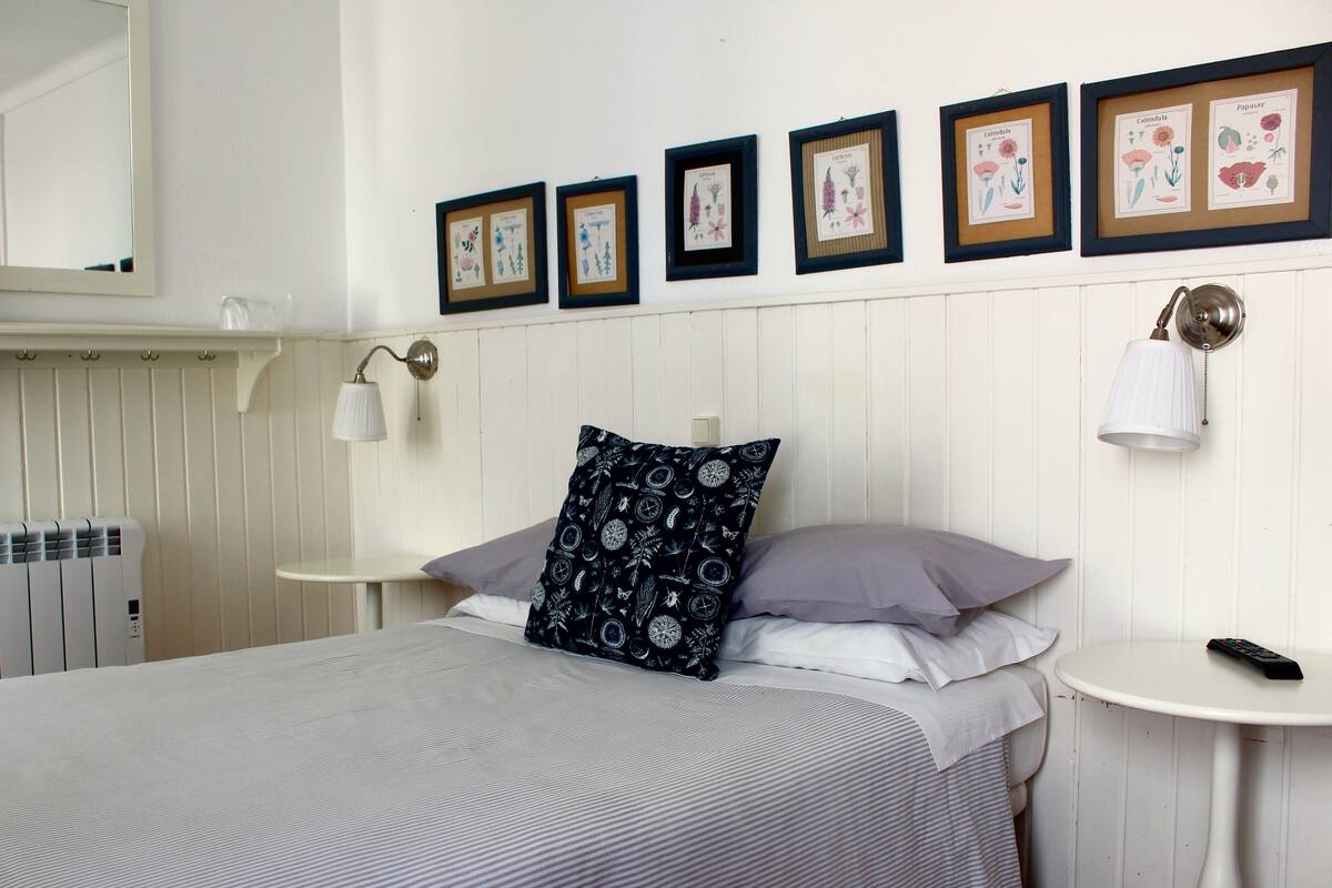 Un dormitorio ordenado con una cama, una mesita de noche, una lámpara de pared e ilustraciones botánicas enmarcadas.