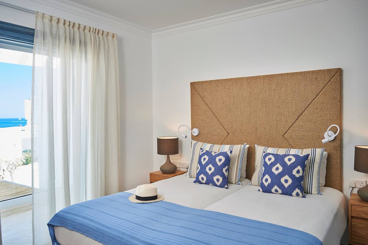 Een net opgemaakt bed met een blauwe spreideken, twee kussens met een patroon, een witte hoed en een houten hoofdbord.