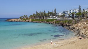 A beach with clear turquoise water, a rocky shoreline, and a row of white buildings with balconies overlooking the sea. - Nausicaa Luxury Villas By Louis Hotels (Paralimni)