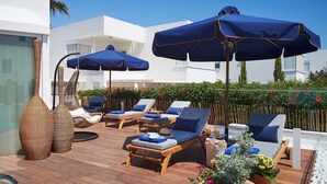 A sunlit outdoor lounge area with wooden deck, lounge chairs, and blue umbrellas. - Nausicaa Luxury Villas By Louis Hotels (Paralimni)