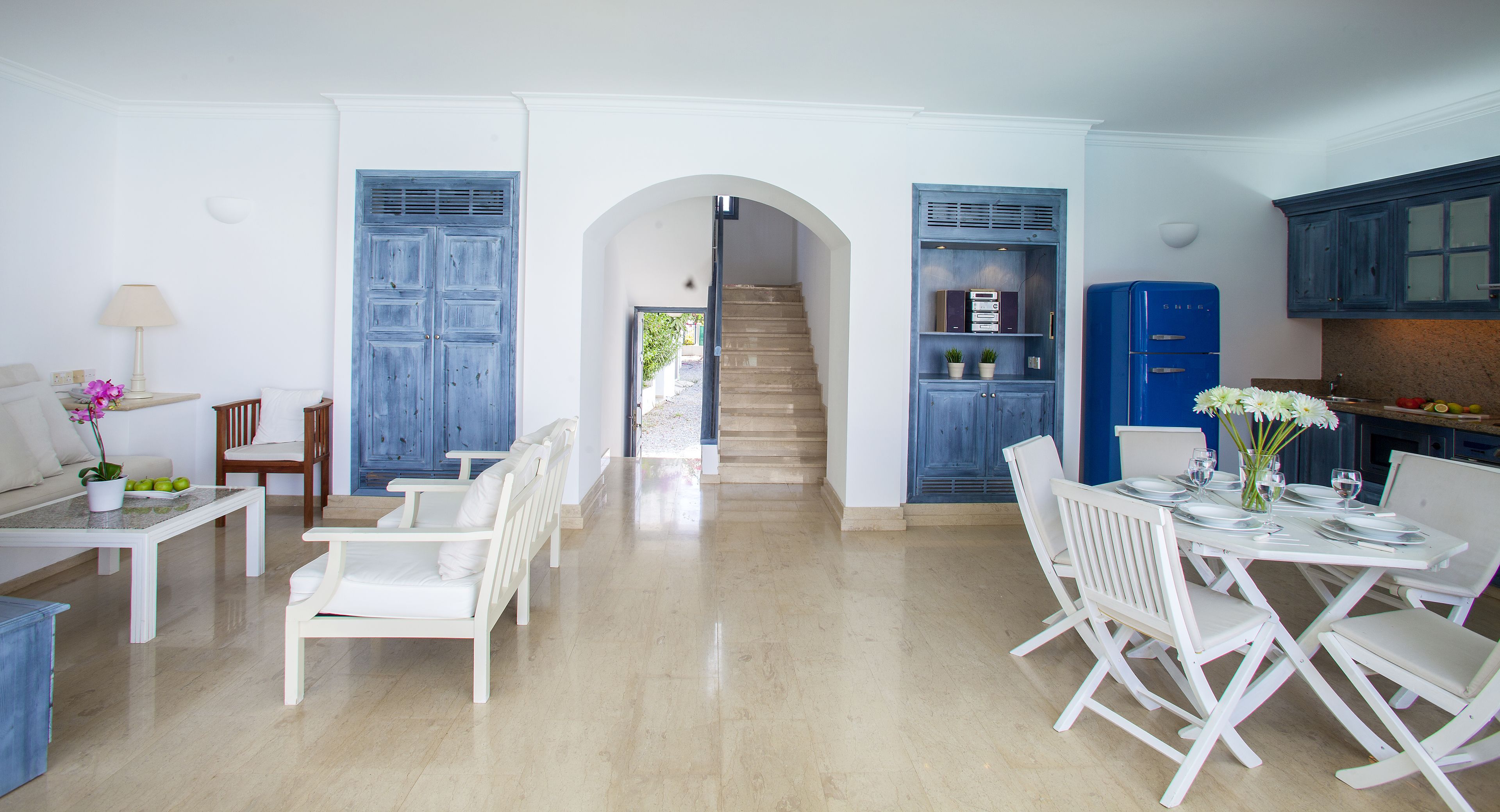 Un amplio salón con una mesa de comedor lista para cuatro comensales, muebles blancos y una escalera que sube a otro nivel.