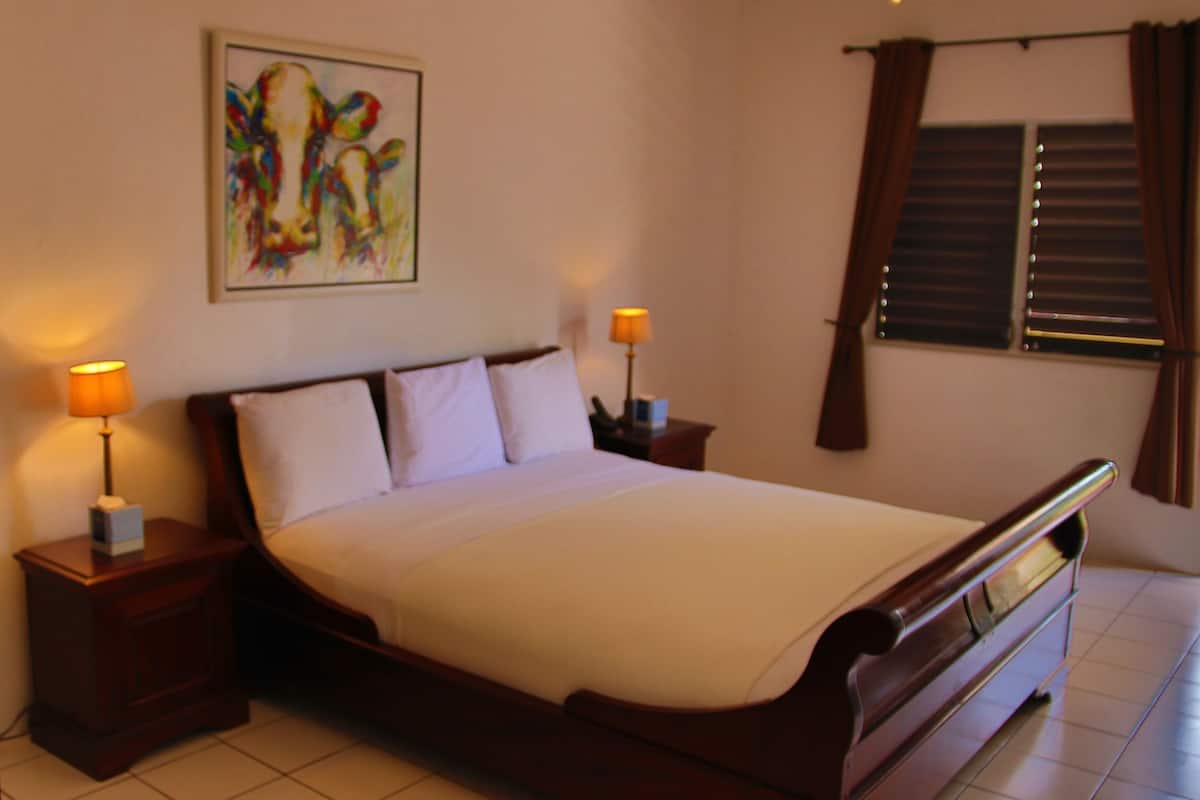 A hotel room with a large bed, two bedside lamps, a nightstand, a wooden headboard, a window with blinds, and a colorful abstract painting on the wall.