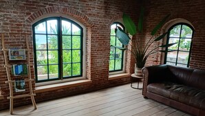 Een kamer met een bakstenen muur, houten vloer, een bruine leren bank en twee grote rondboogvensters met uitzicht op groen.