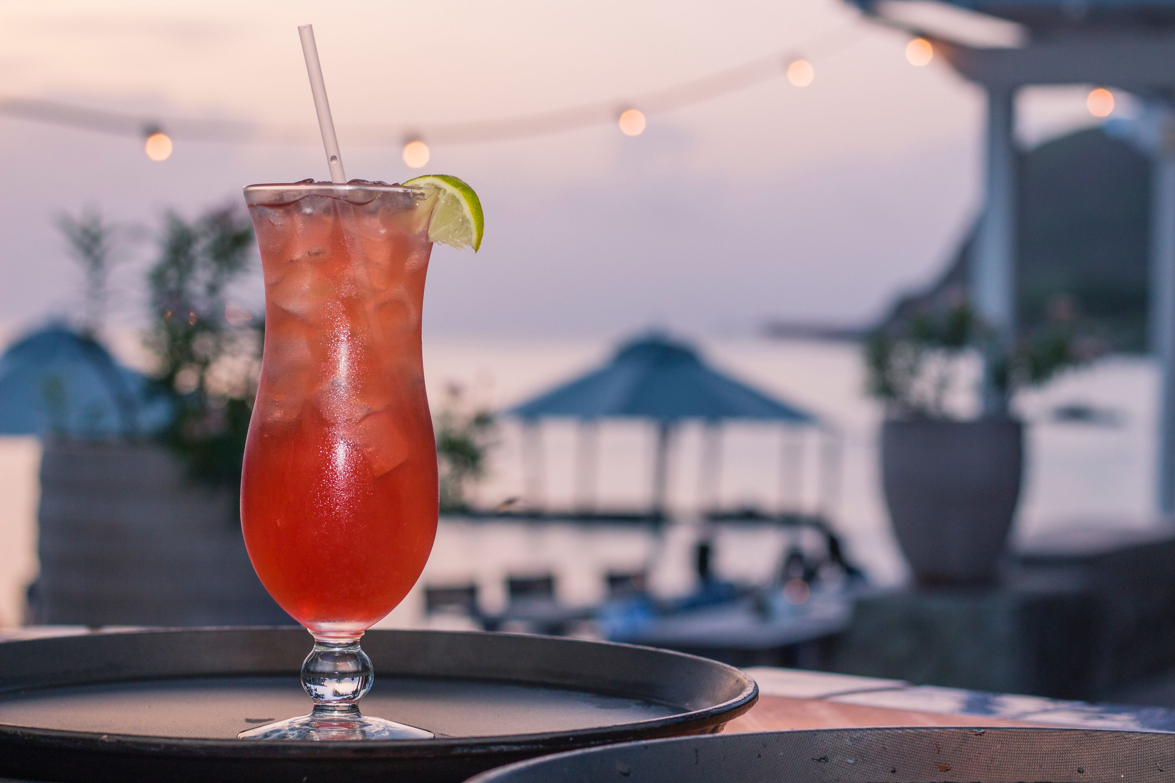 A tall glass with a straw and a lime wedge on a tray, set against a blurred background with string lights.