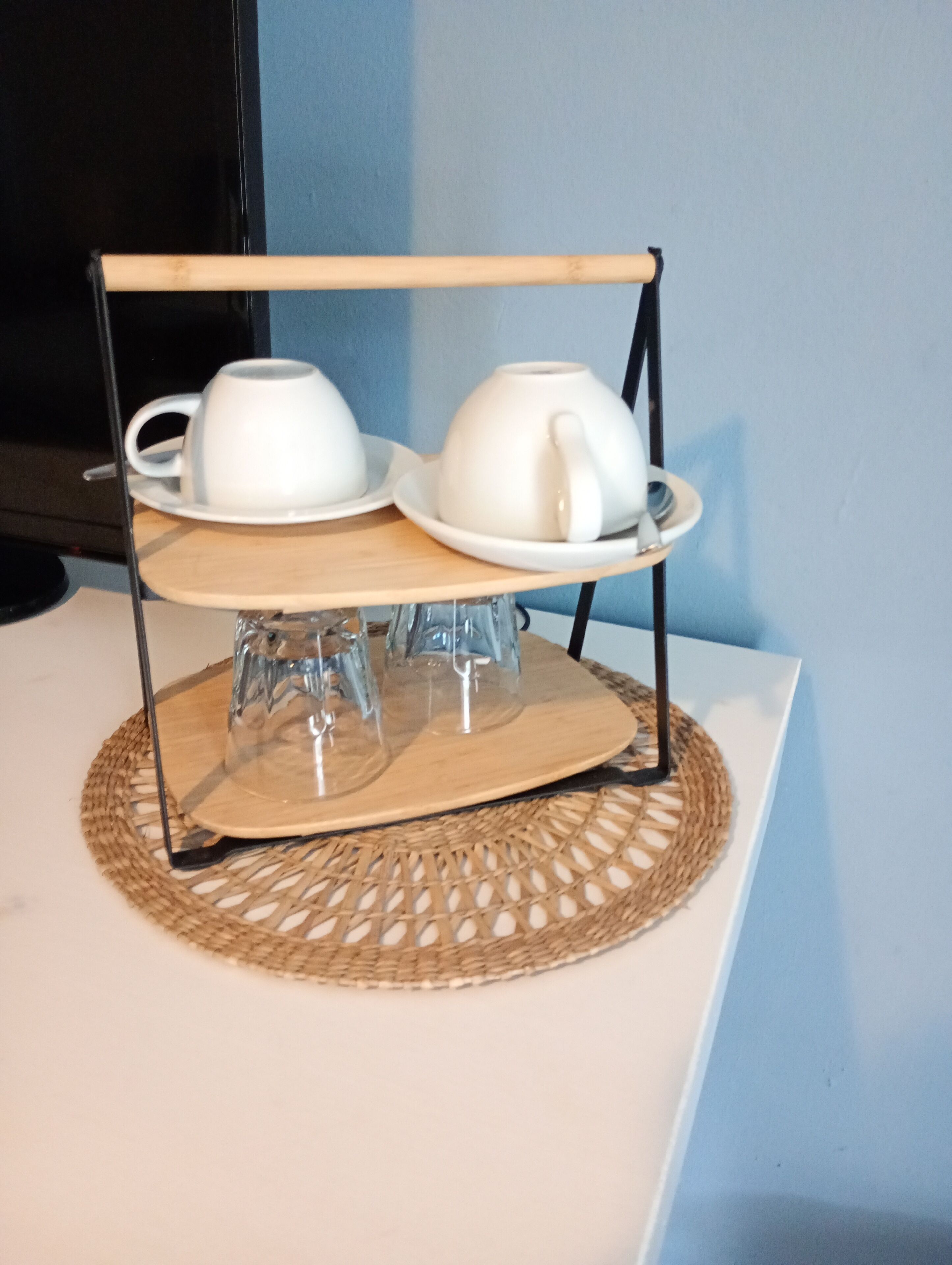 A two-tiered white ceramic tea set on a woven placemat, displayed on a white table against a blue wall.