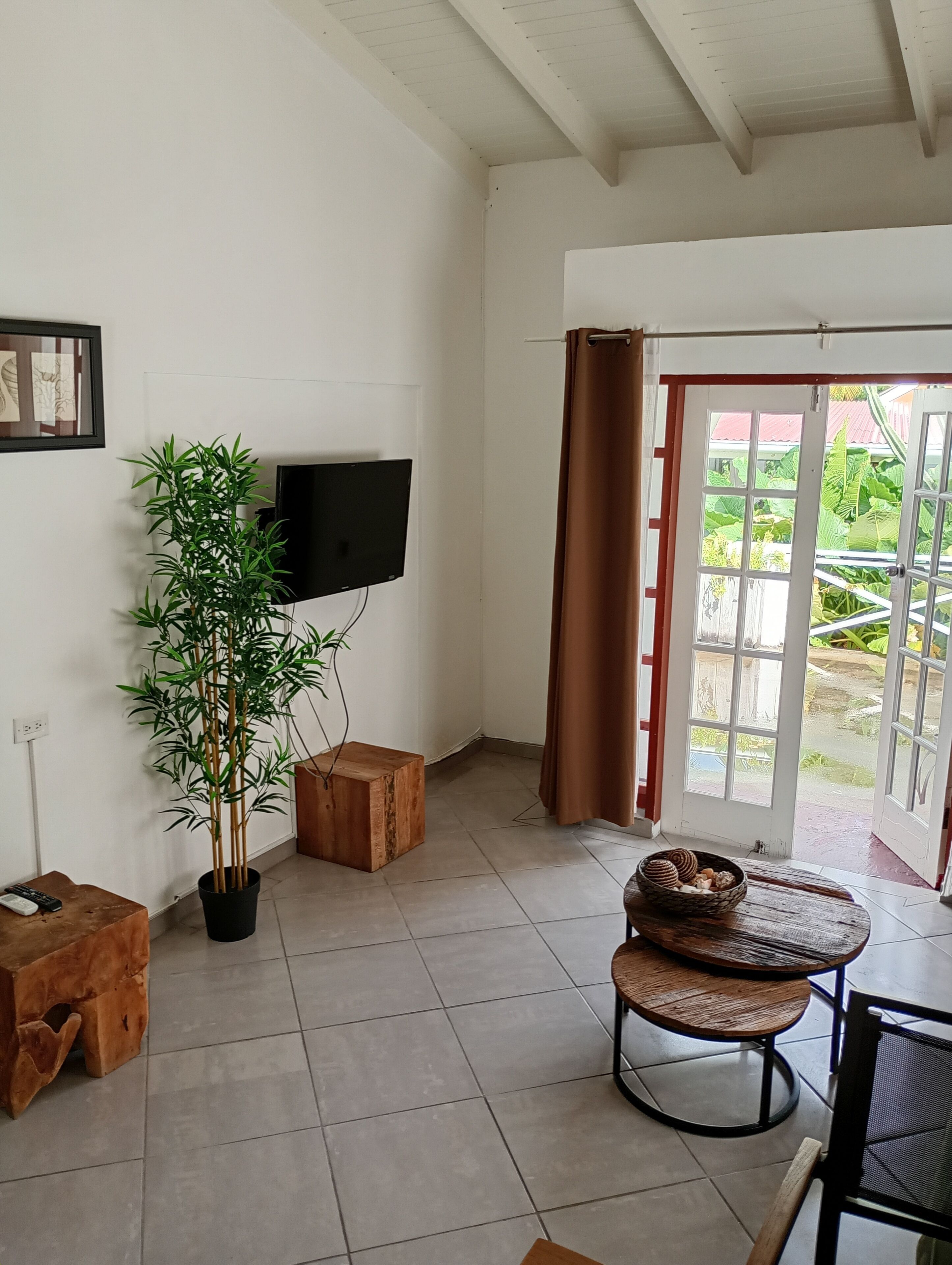 Una sala de estar con televisor, una planta de bambú en maceta, un taburete de madera, una mesa redonda de madera con una cesta tejida y una puerta corrediza de vidrio que da a un área exterior.