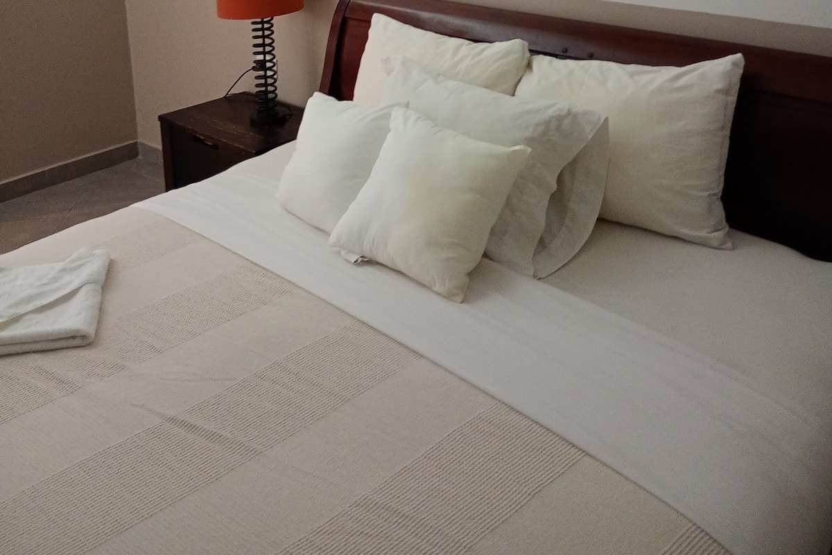 A neatly made bed with white linens and pillows, a wooden headboard, a small bedside table with a lamp, and a framed picture on the wall.