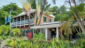 Een tweelaags gebouw met een balkon, omringd door weelderige tropische planten en een bordje voor 'The Old Sun House Restaurant Bar'.