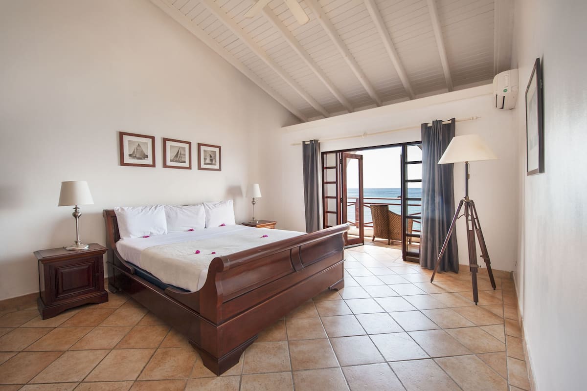 A bedroom with a large bed, two bedside tables, a lamp, a tripod, and a view of the sea through an open door.