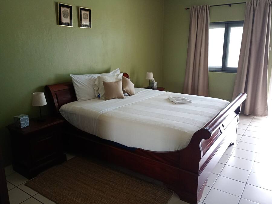 A neatly made bed with white bedding, a wooden headboard, and a brown headboard. There are two bedside tables with lamps and a tissue box on one. The room has green walls, a window with curtains, and a tiled floor.