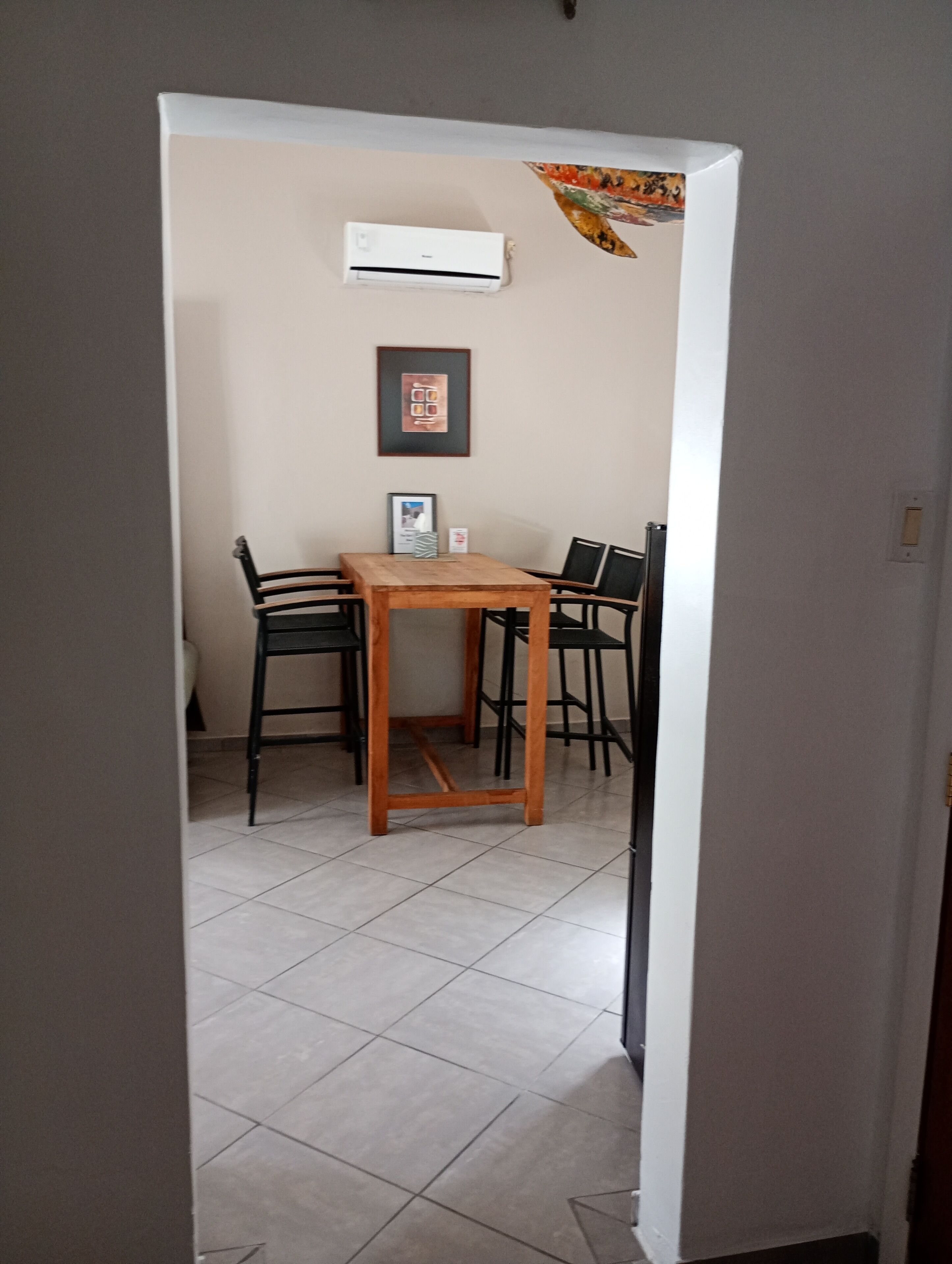 A room with a tiled floor, a wooden table with four chairs, an air conditioning unit, and a framed picture on the wall.