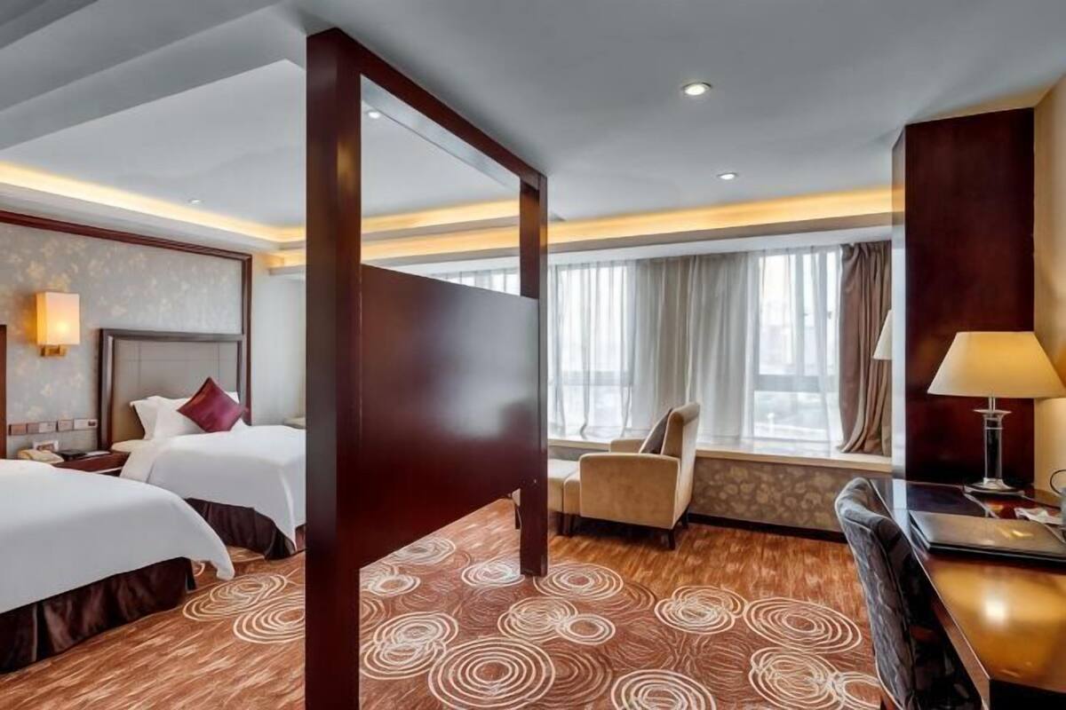 Une chambre d’hôtel avec deux lits, un bureau, une chaise, une grande fenêtre avec des rideaux et une moquette à motifs.