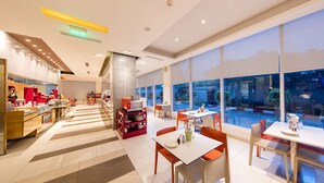 A modern restaurant with a central serving area, multiple dining tables, and large windows offering views of the outdoors. - ibis Chennai SIPCOT Hotel (Tirupporur)