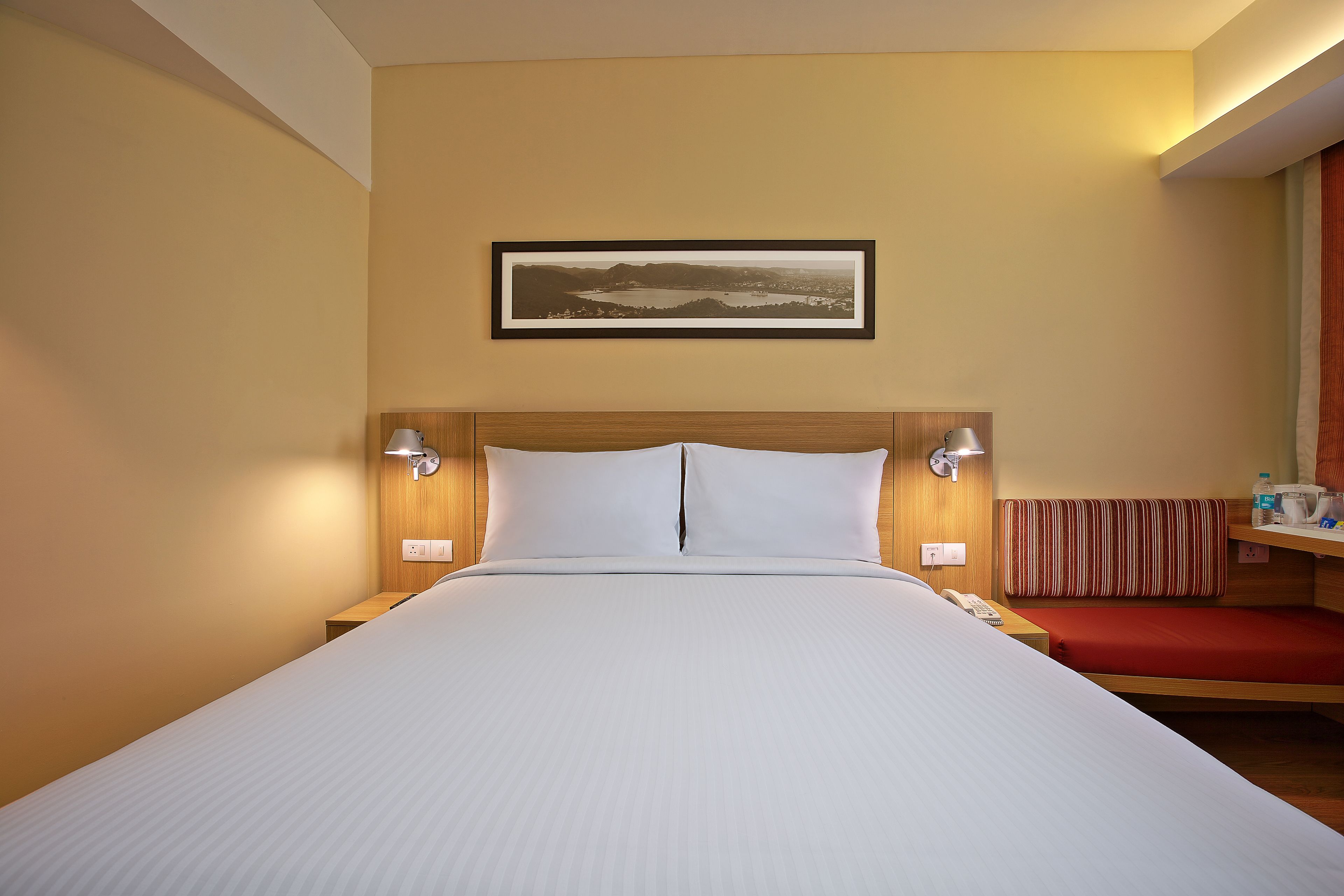 Ein Hotelzimmer mit einem großen Bett, einem hölzernen Kopfteil, einem Nachttisch mit Lampe und einer kleinen Sitzecke mit rotem Kissen.