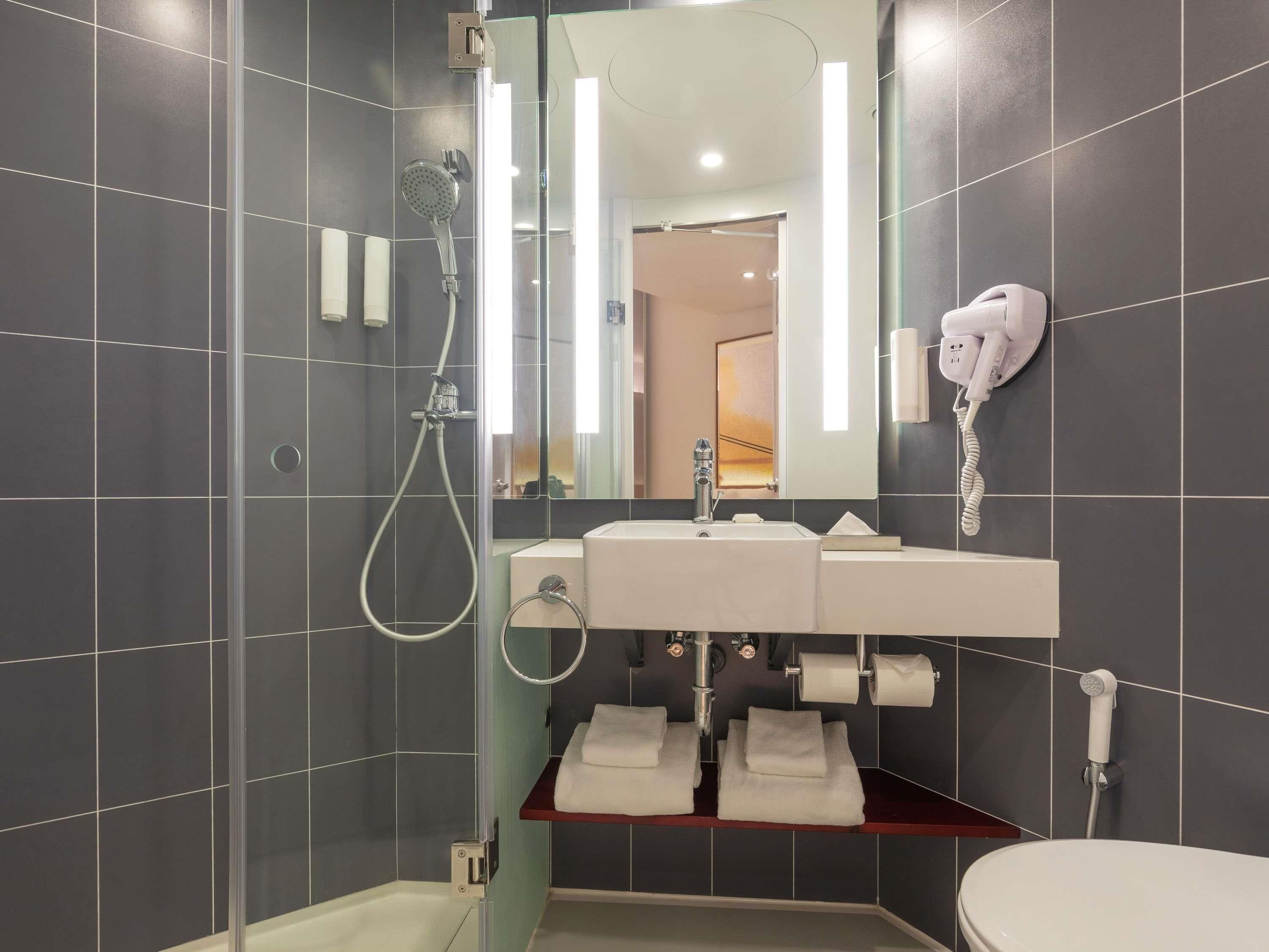 Une salle de bain moderne avec une douche à l’italienne en verre, un lavabo blanc avec un miroir au-dessus et un sèche-cheveux fixé au mur.