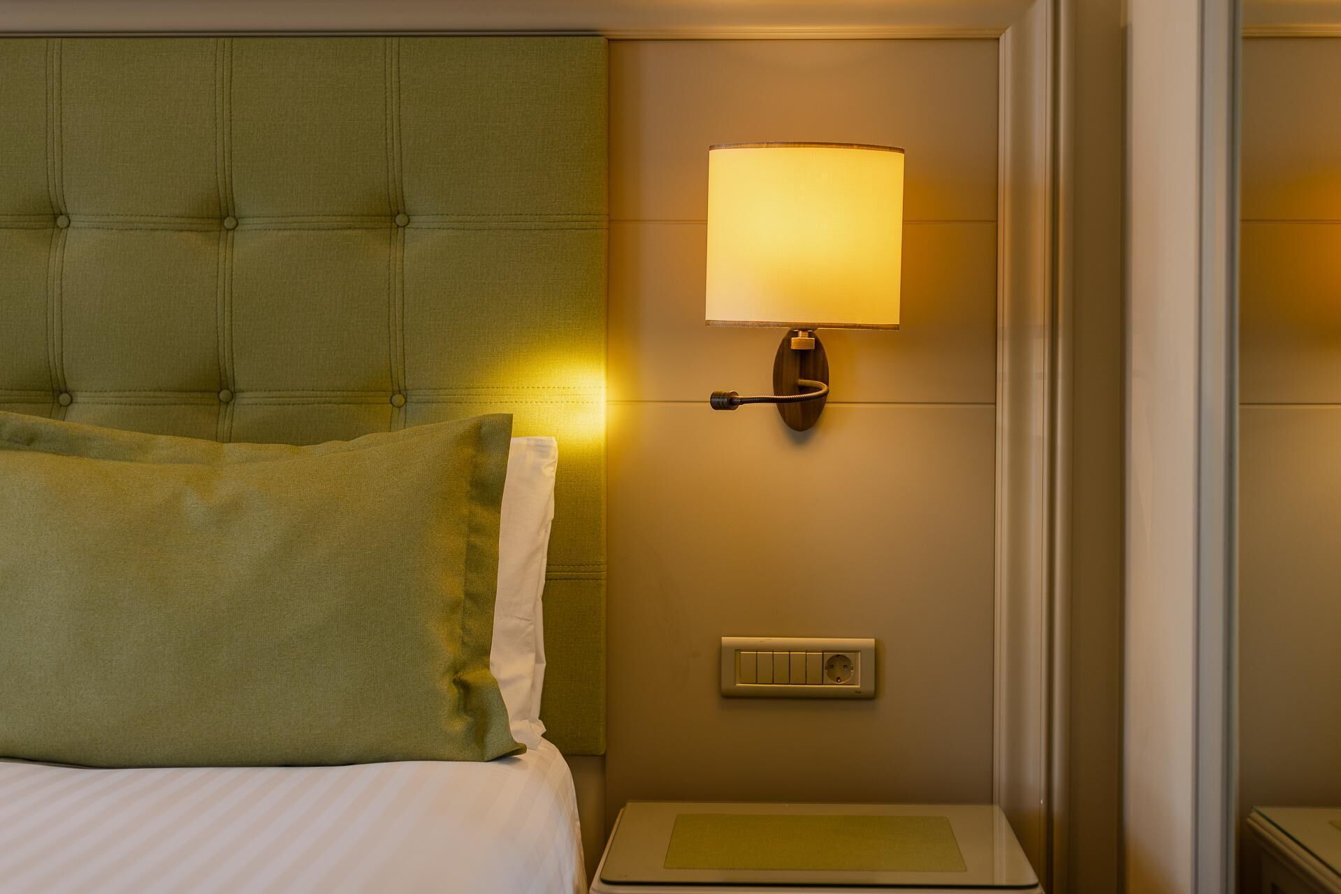 Une chambre d’hôtel avec une tête de lit recouverte d’un tissu vert, une lampe de chevet, une table de chevet et une prise de courant.