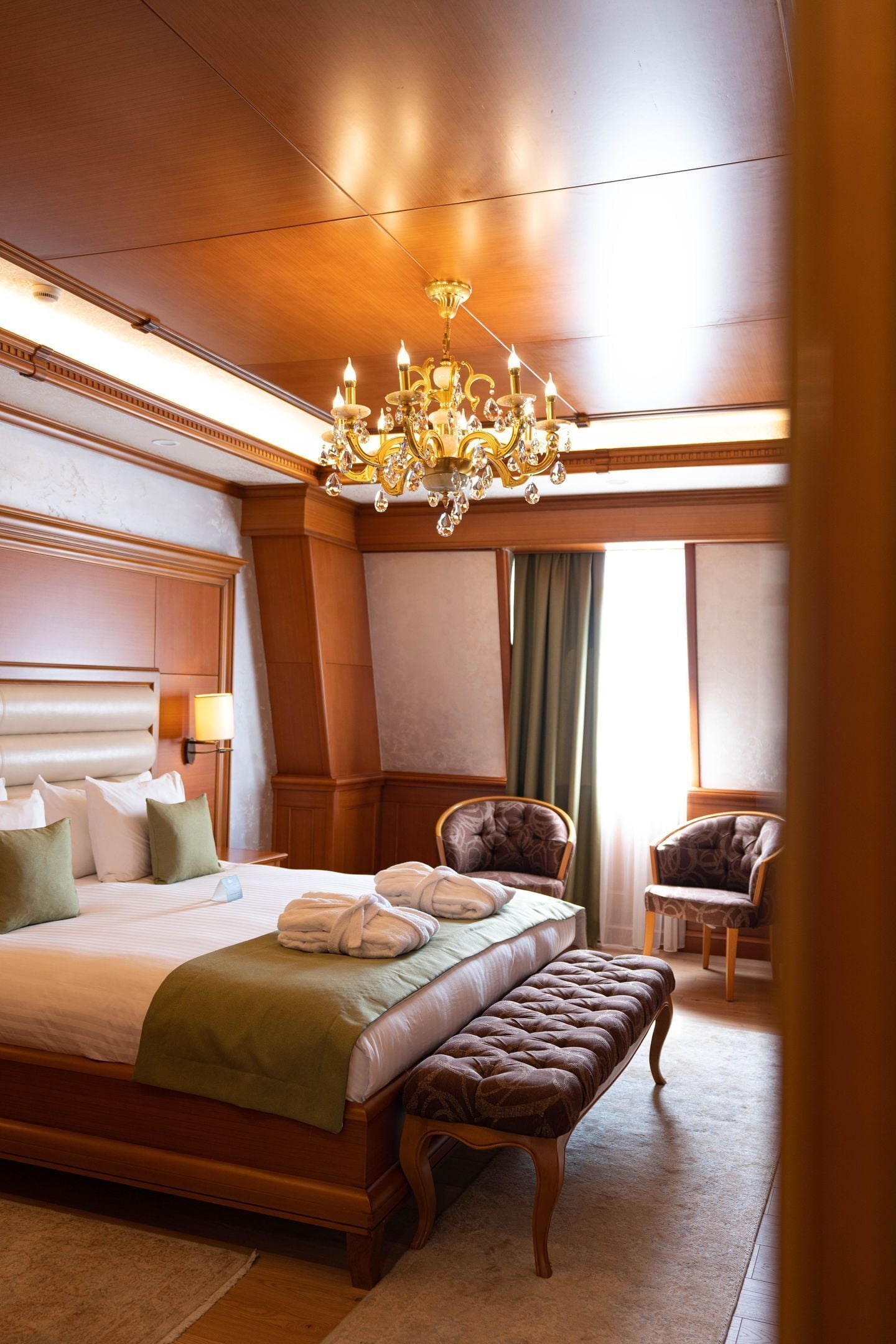 A hotel room with a large bed, a bench, two armchairs, a chandelier, and a window with curtains.
