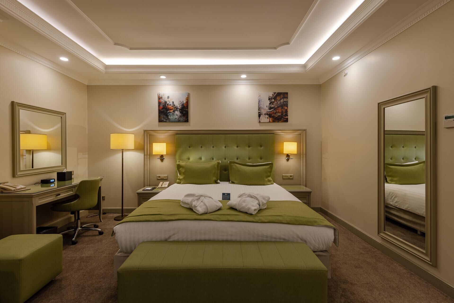 A hotel room with a large bed, two bedside lamps, a desk with a chair, a mirror, and artwork on the walls.