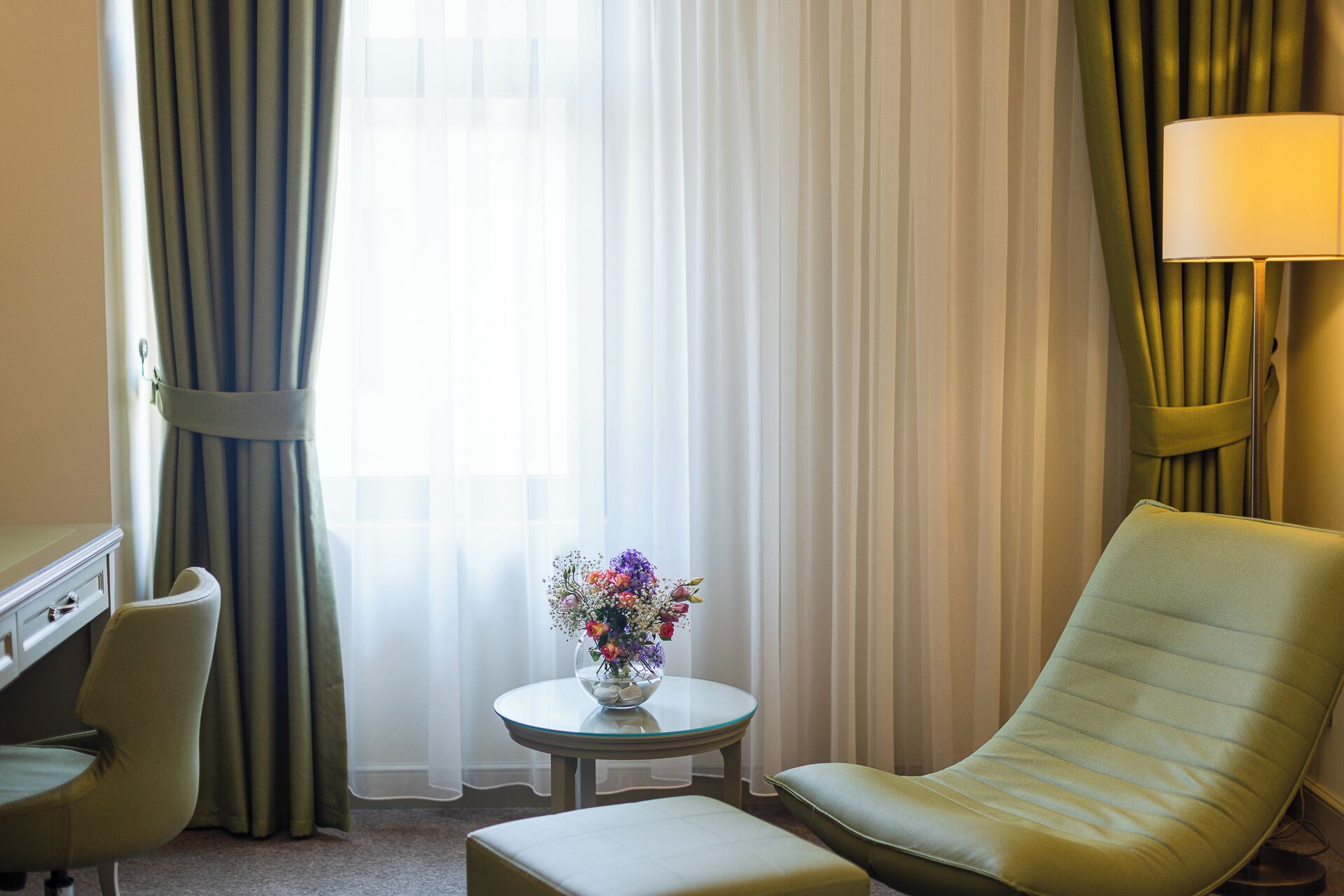 Una habitación de hotel con un sillón verde, una mesita redonda con un florero y una lámpara, además de un escritorio.