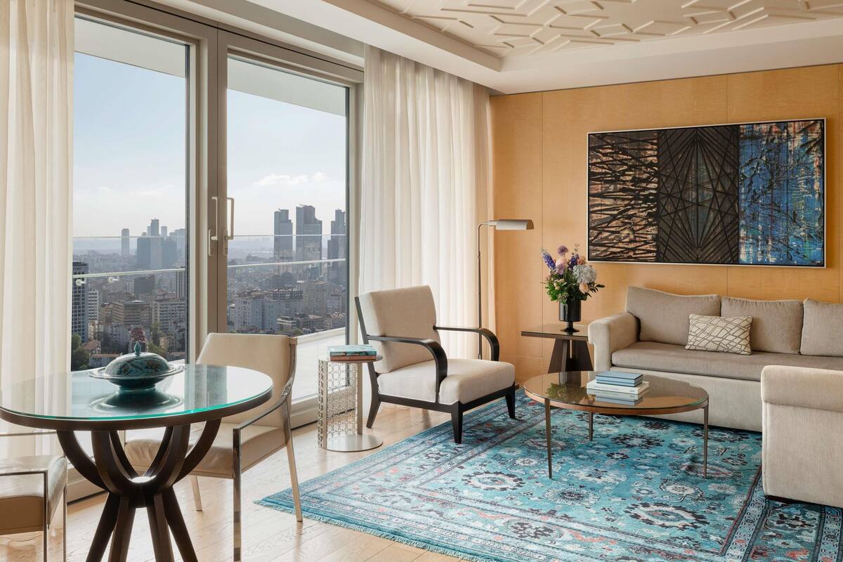 A modern living room with a sofa, armchairs, a glass coffee table, a patterned rug, and a city view through large windows.