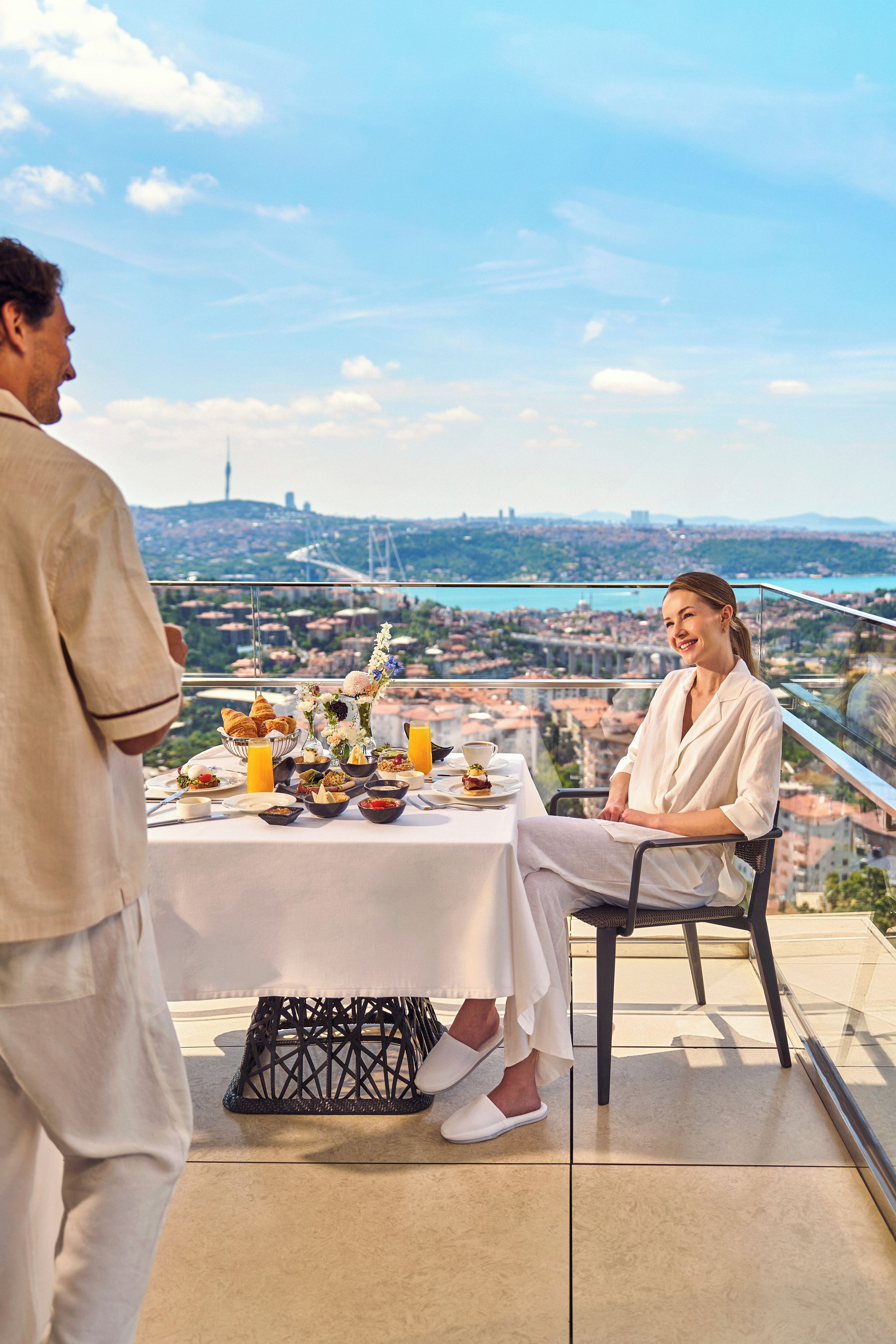 A dining table with a city view, a man and a woman seated, and a table set with food and drinks.