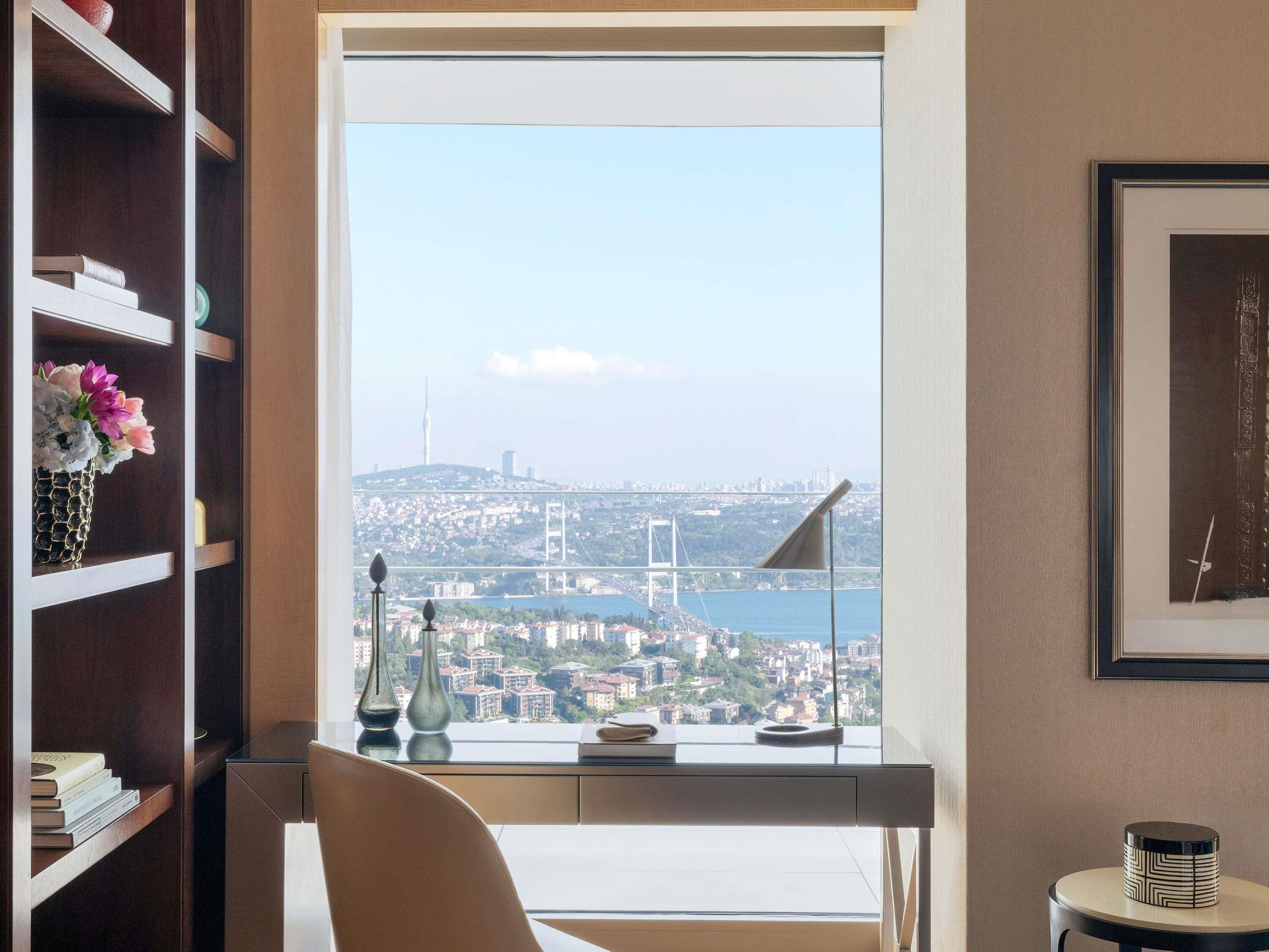 Une pièce avec une grande fenêtre, un bureau, une chaise, une bibliothèque et un tableau encadré au mur.