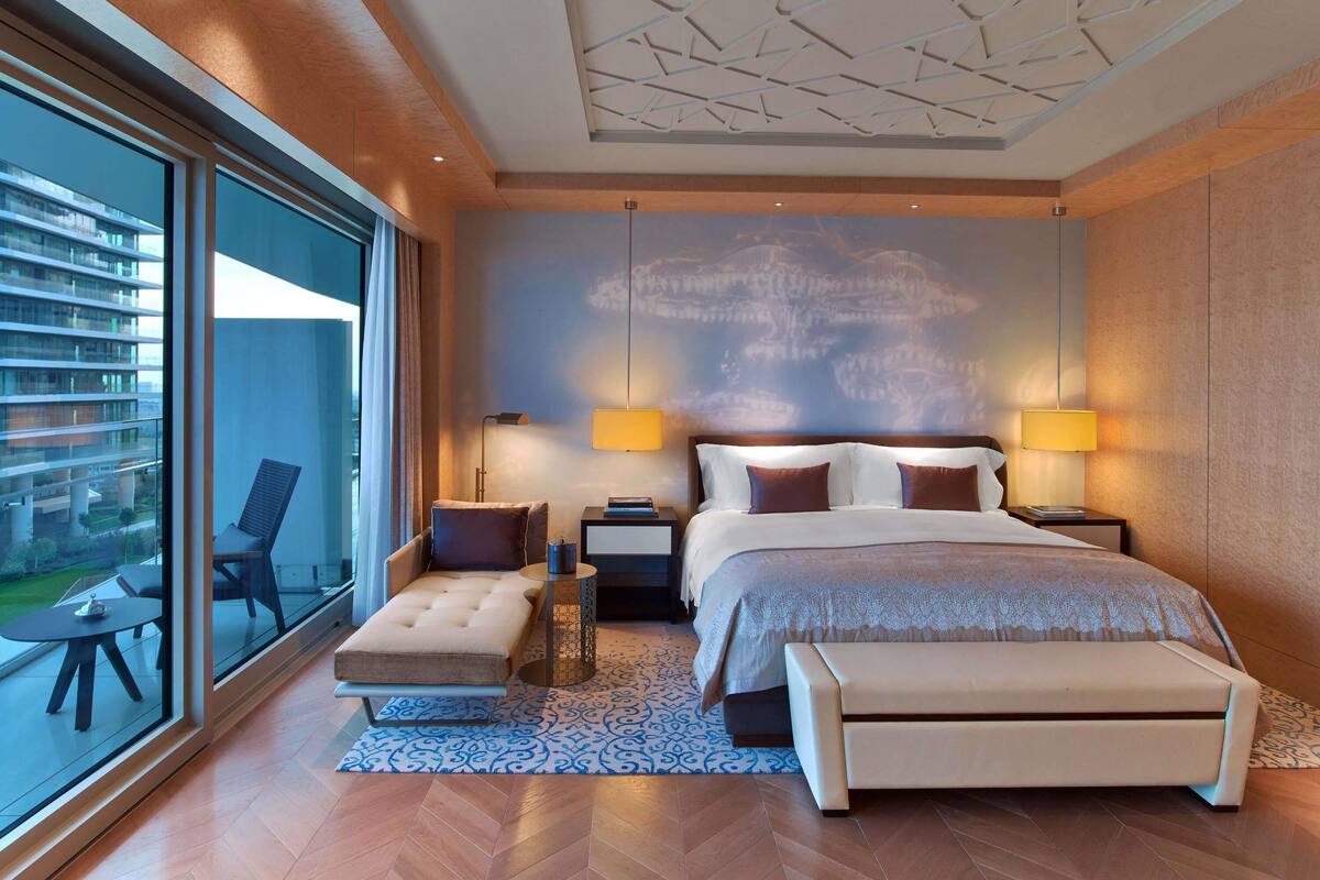 A modern bedroom with a large bed, two bedside lamps, a bench, and a view of a building through a large window.