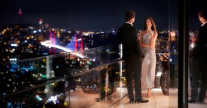 A couple standing on a balcony overlooking a cityscape at night.