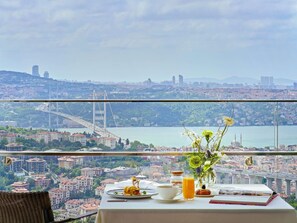 A table with breakfast items, a vase with flowers, and a view of a cityscape with a bridge.