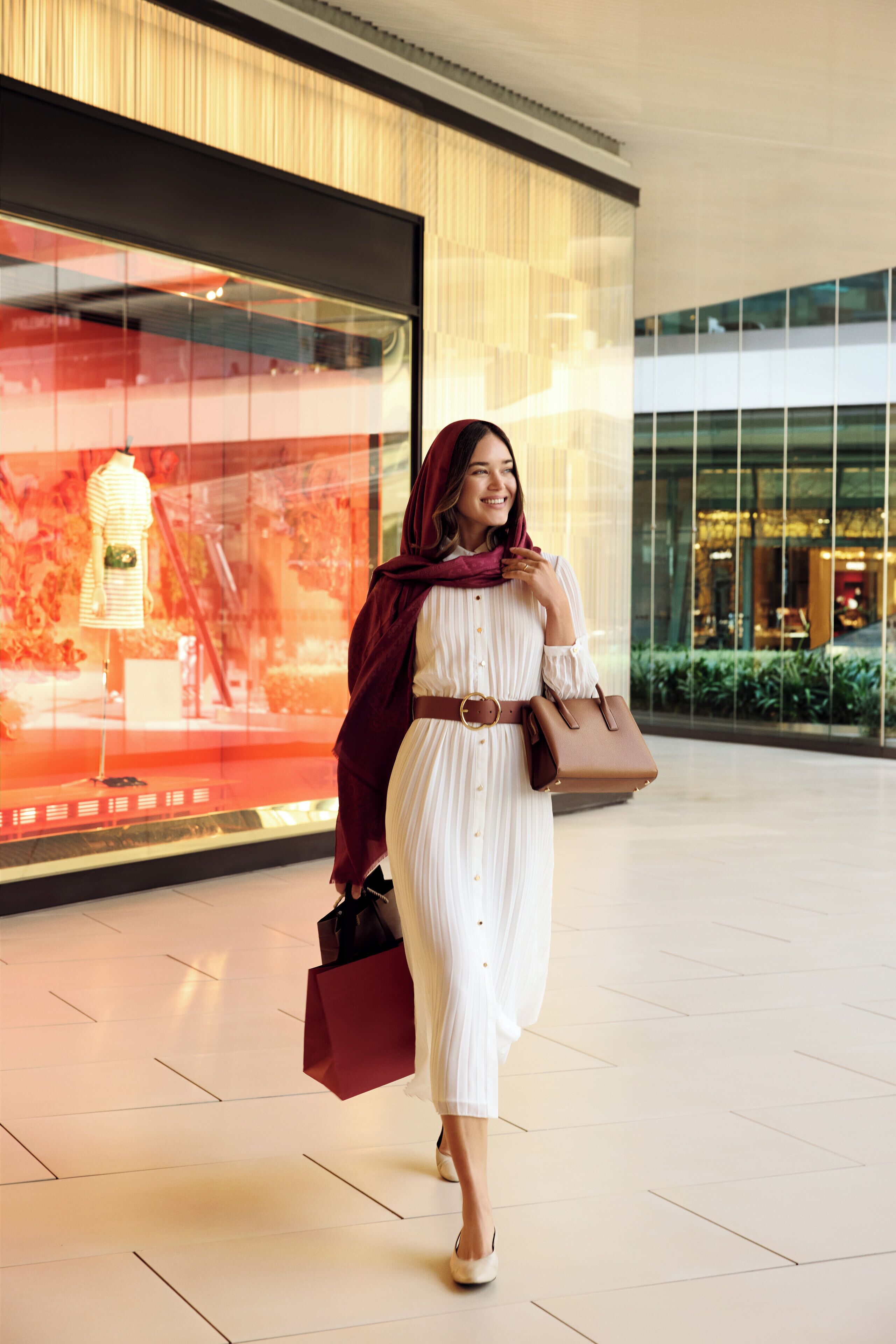A woman in a white dress with a brown belt and a brown handbag walking in a shopping mall.
