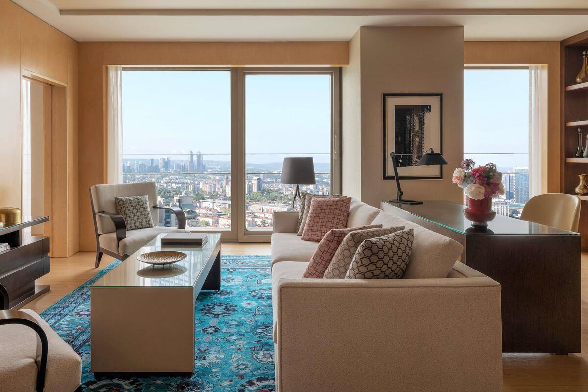 A modern living room with a city view, a glass coffee table, a sofa, a chair, and a desk with a lamp.