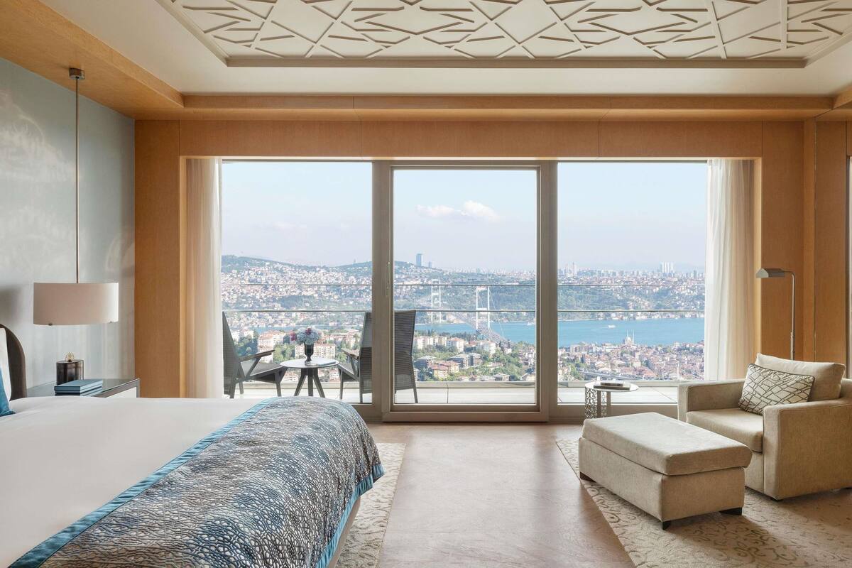 A hotel room with a large bed, a sofa, a chair, and a view of a cityscape through large windows.